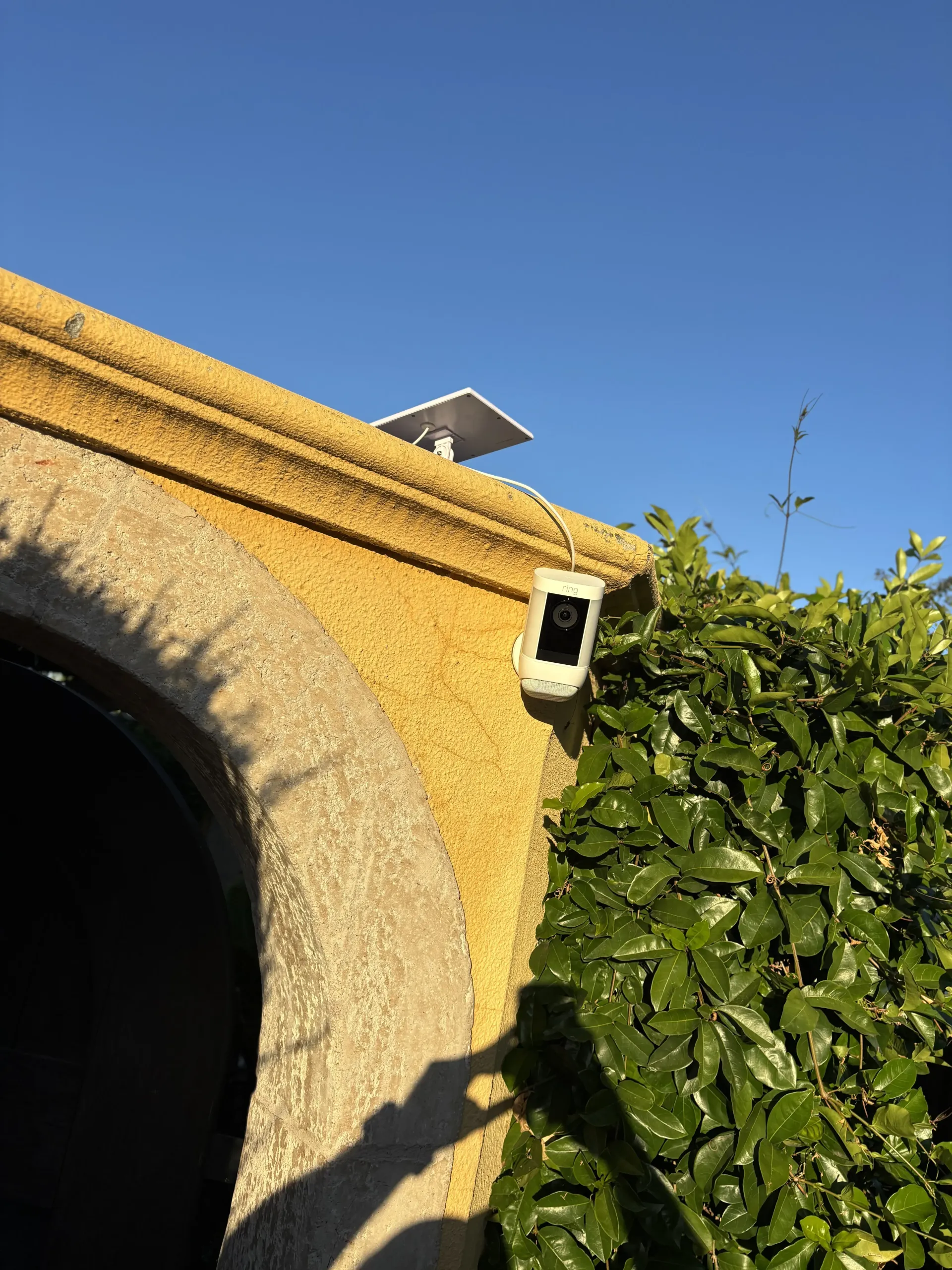 A security camera mounted on a textured yellow stucco wall with a small solar panel on the roof ledge above.