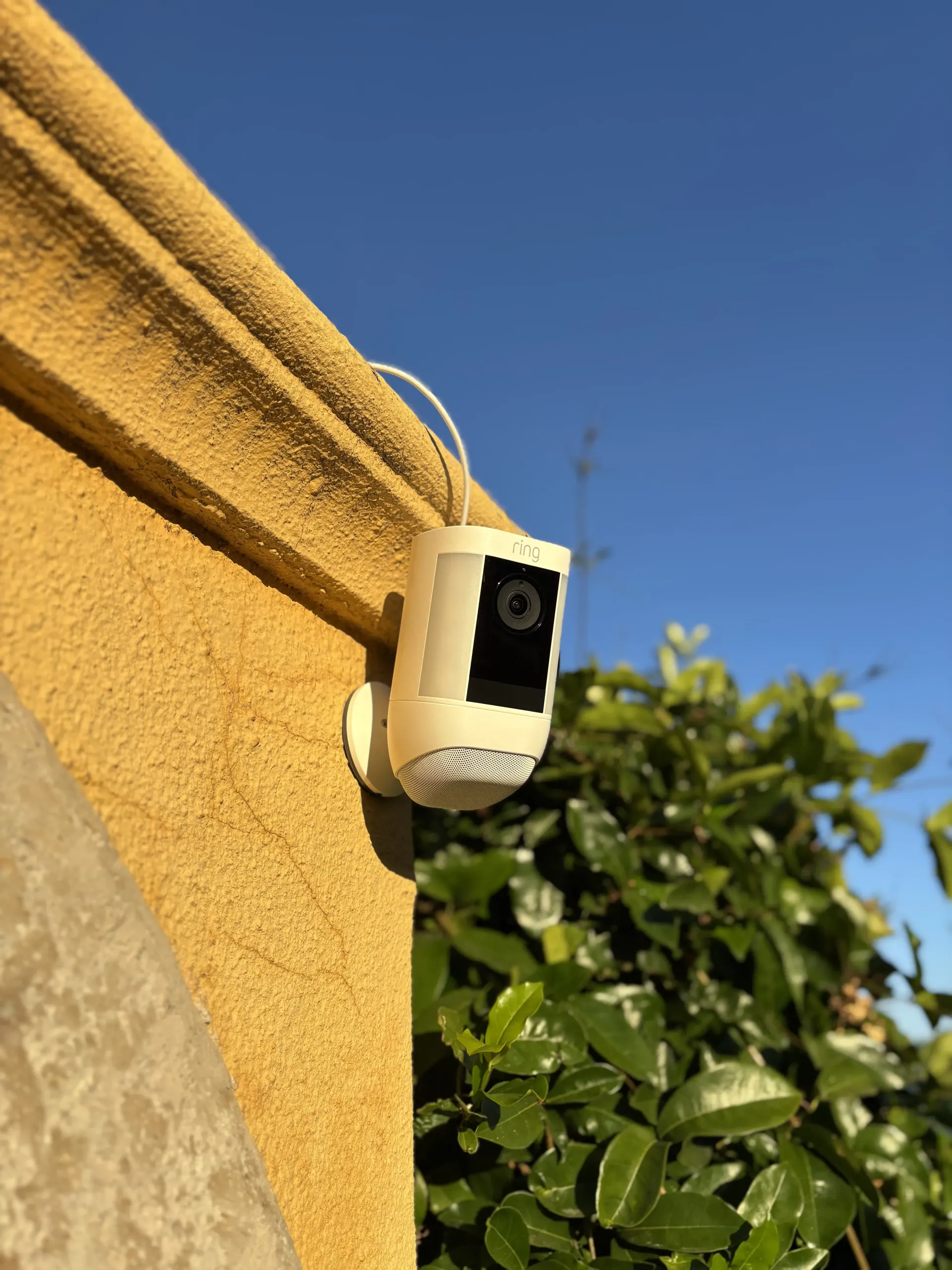 A white security camera is mounted on a yellow stucco wall outdoors with a green leafy bush and blue sky in the background.