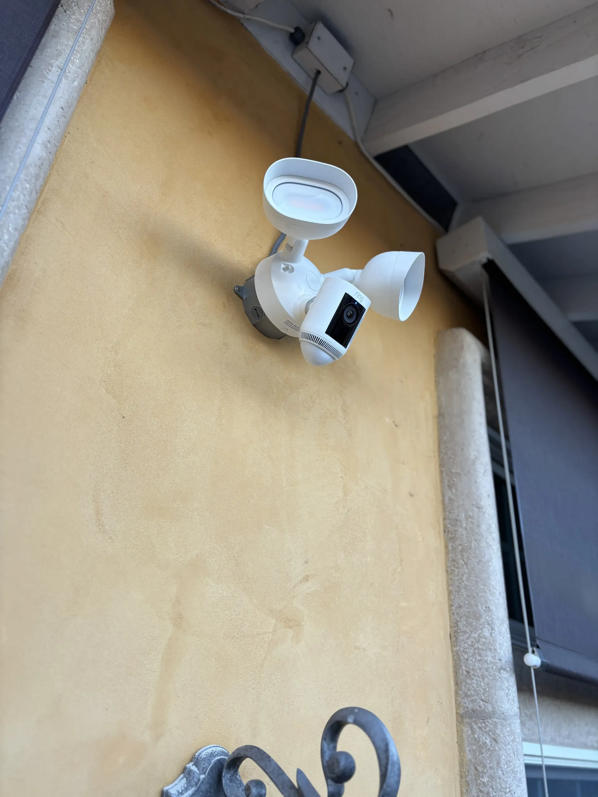 A white smart security camera with dual LED floodlights mounted on a yellow textured exterior wall.
