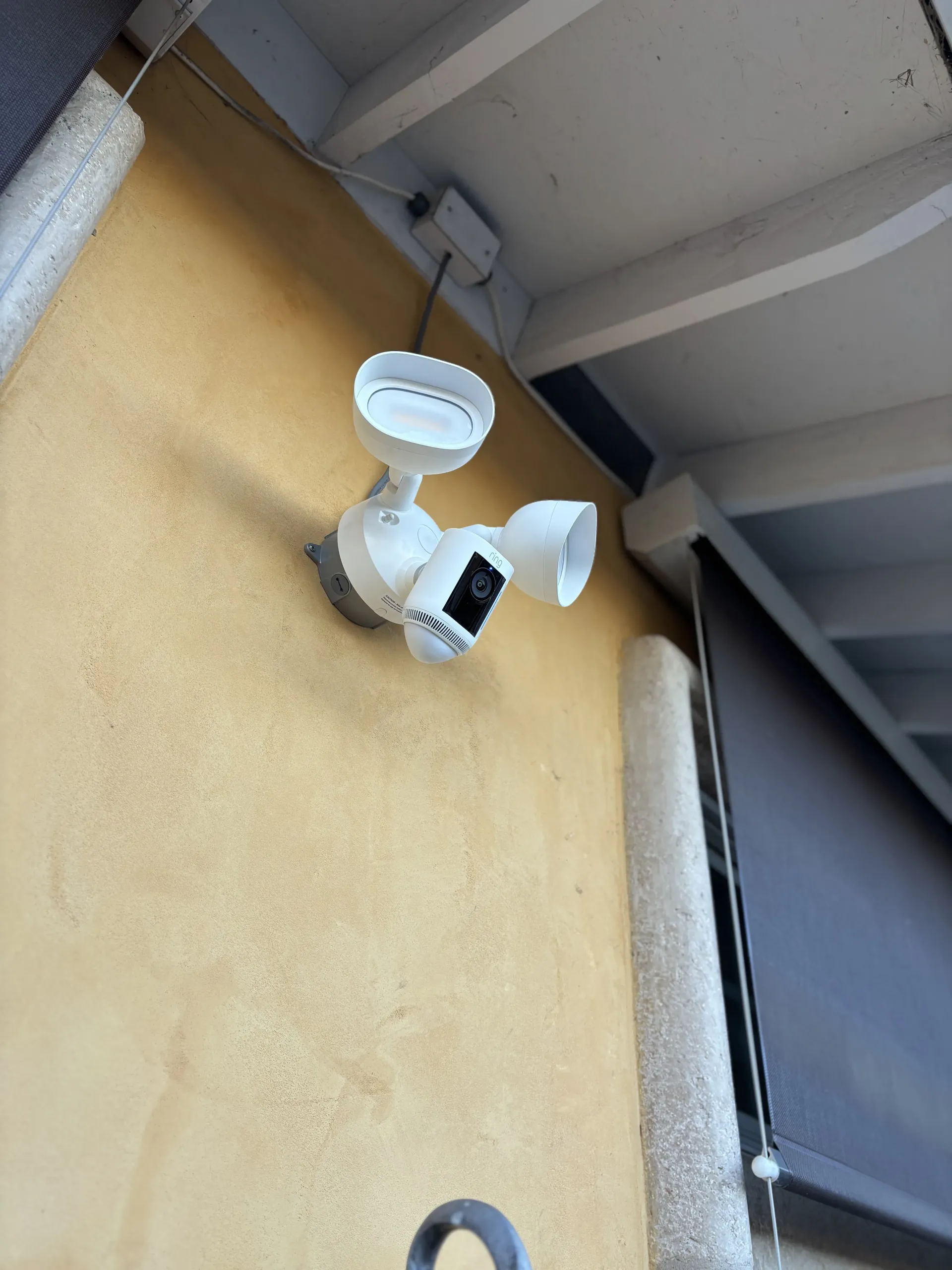 A white security floodlight camera mounted on a textured, light yellow exterior wall under a porch overhang.