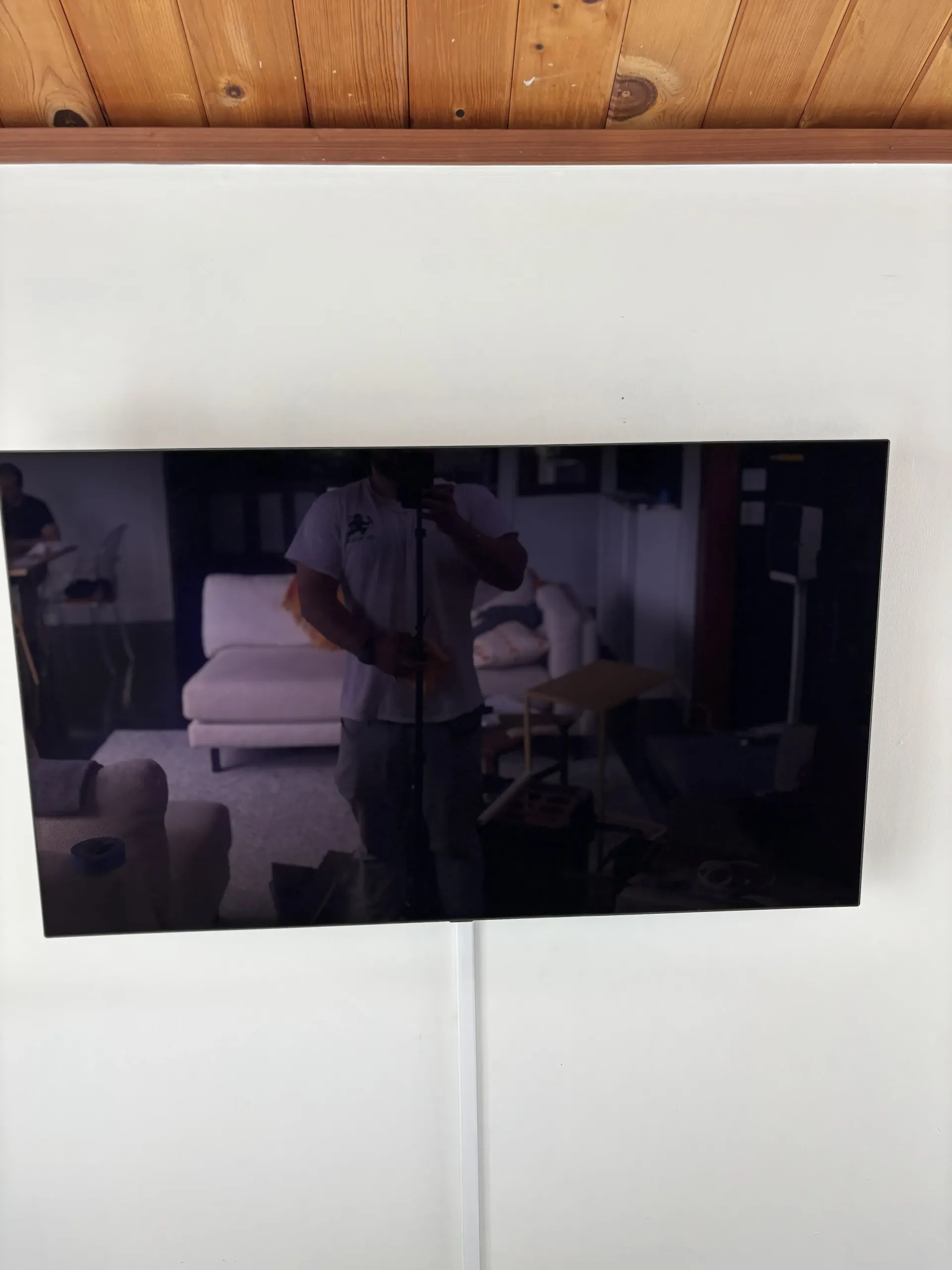 A black television screen mounted on a white wall, showing a reflection of a person standing in a living room.