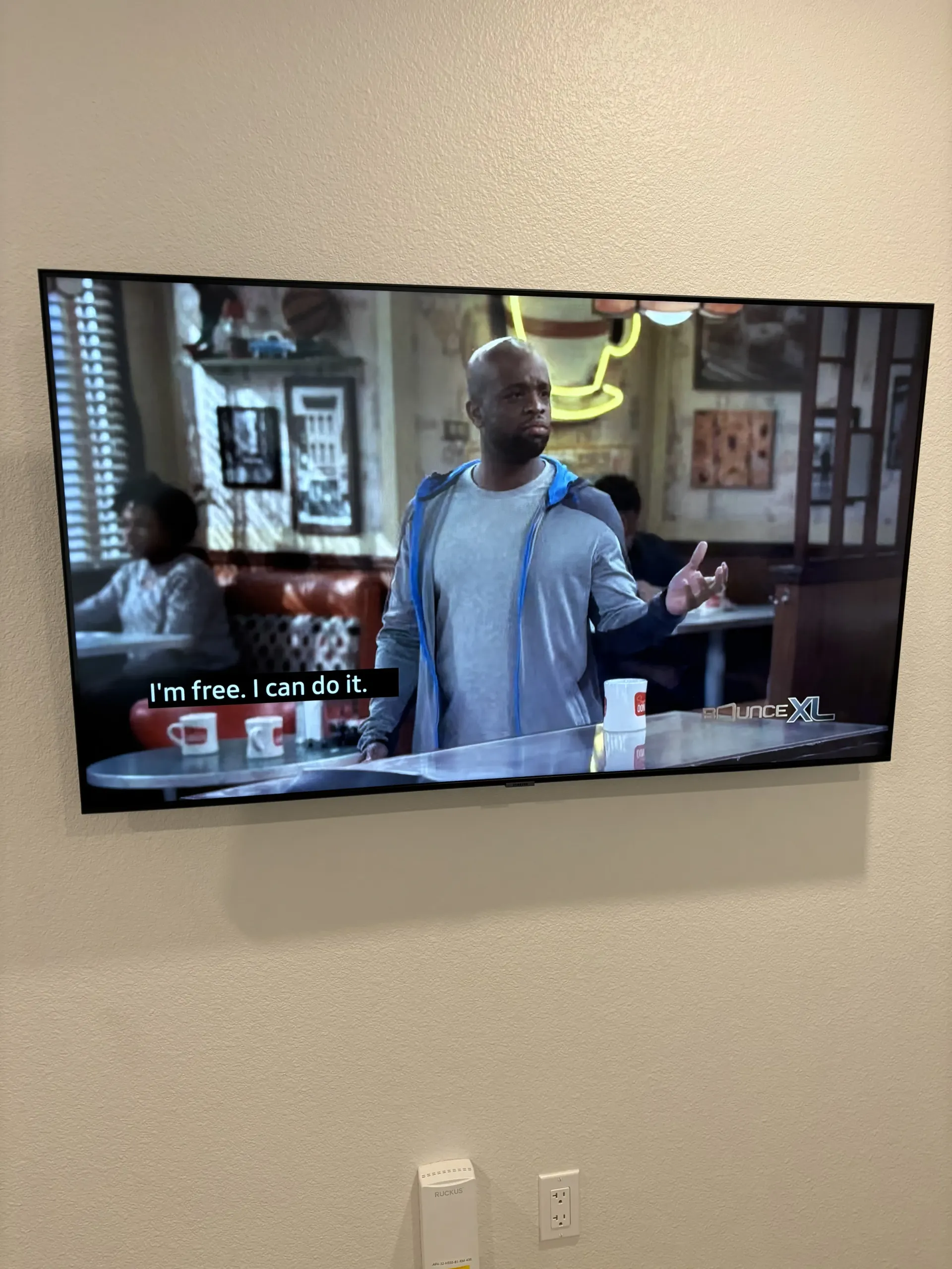 A man in a blue jacket stands in a cafe, speaking while gesturing with his hand, with subtitles on the TV screen.