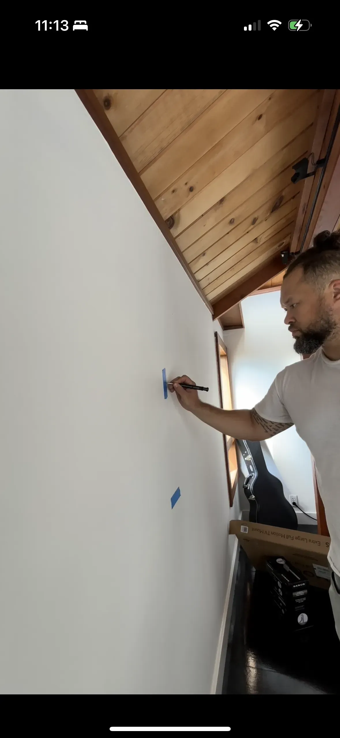 A person marks a spot on a white wall with a pen below a wood-paneled ceiling.