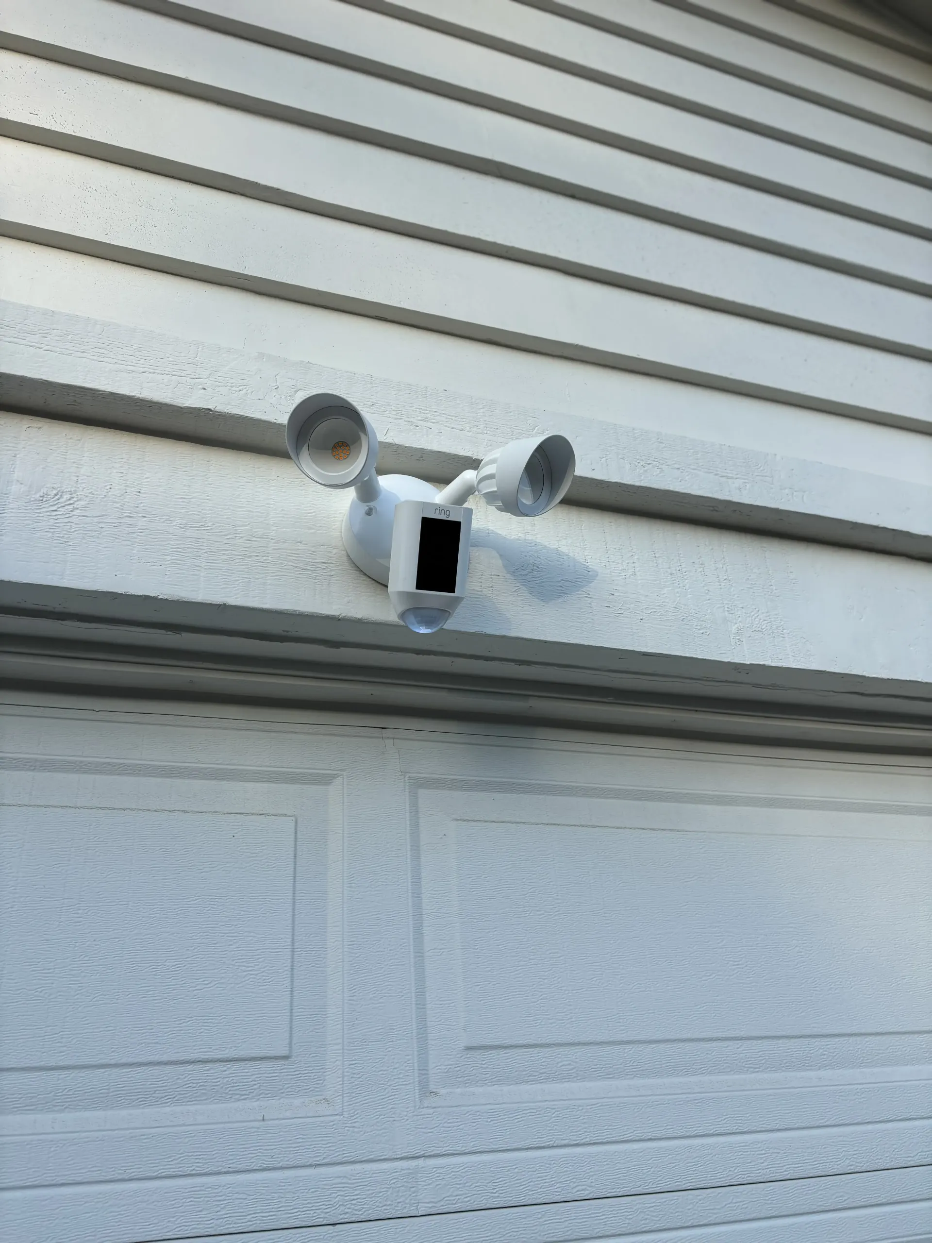 A white dual-head floodlight camera mounted on the siding above a garage door.