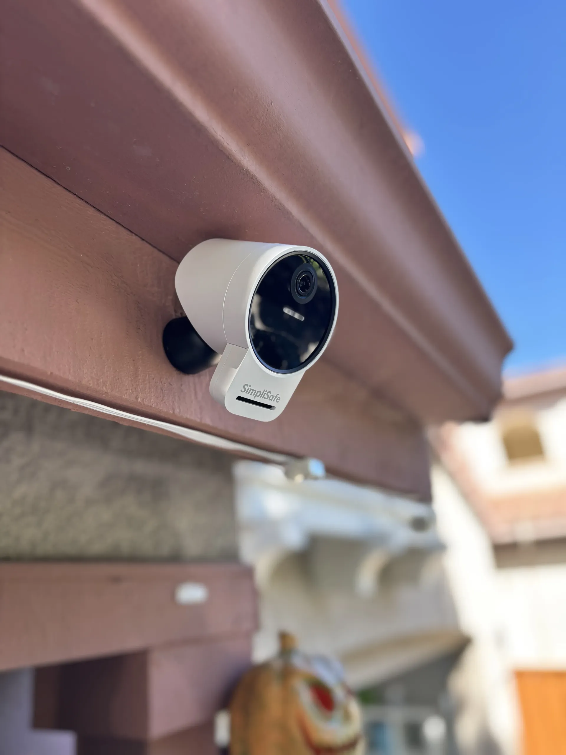 A white smart security camera mounted to a wooden structure outdoors against a bright blue sky.