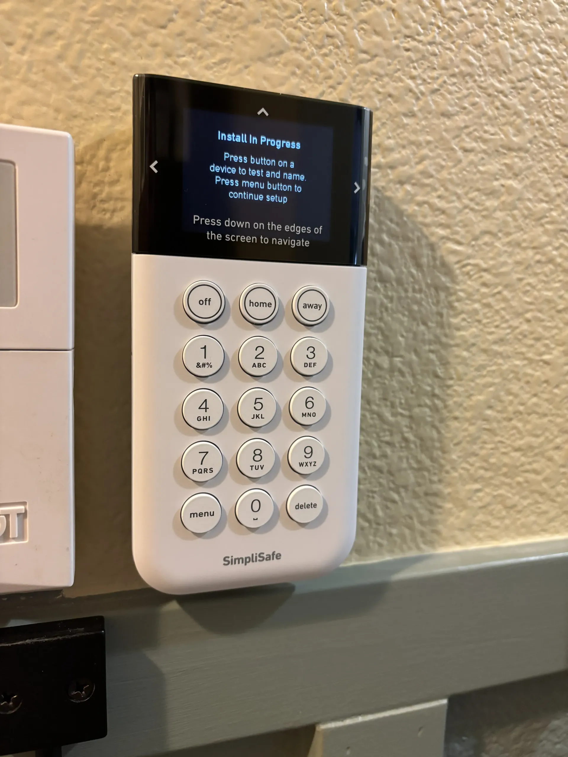 A white SimpliSafe security keypad mounted on a beige wall, displaying an 