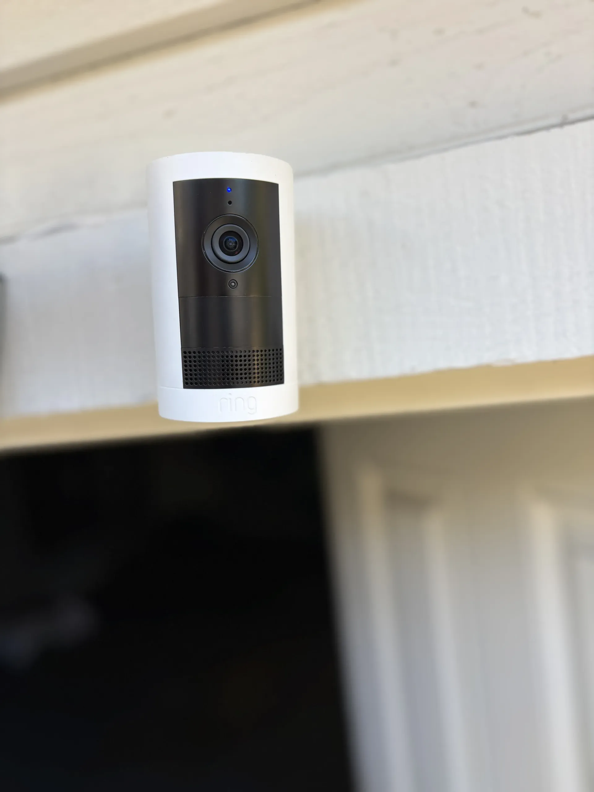 A white wireless security camera mounted above a door frame against a white wall.