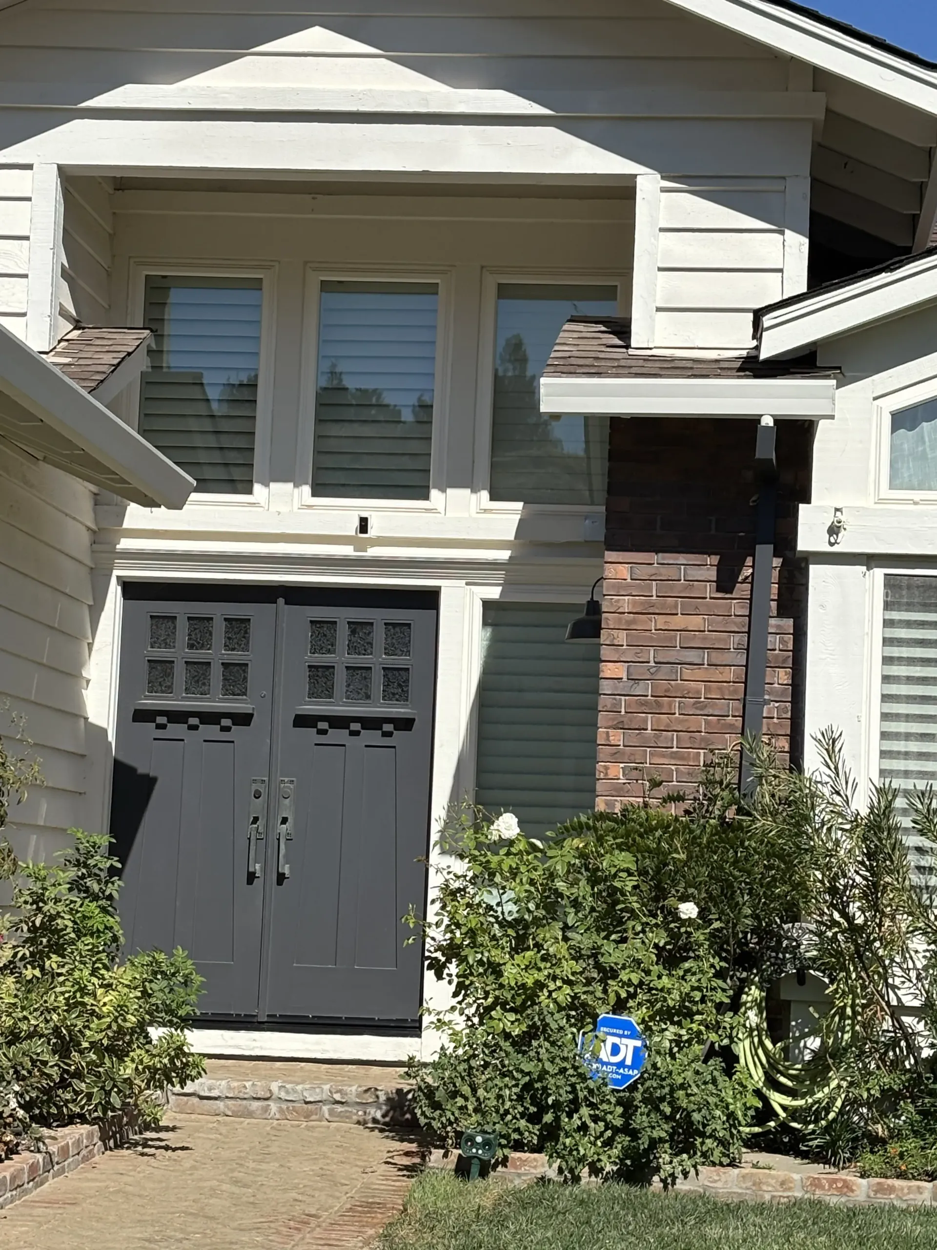 Front entrance of a house with a dark grey double door, brick siding, and a security system sign in the front landscaping.