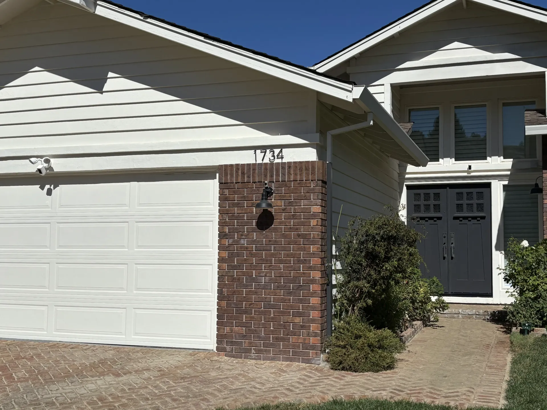 A house with a white garage, brick accent wall, and a dark front door under a gabled roof on a sunny day.