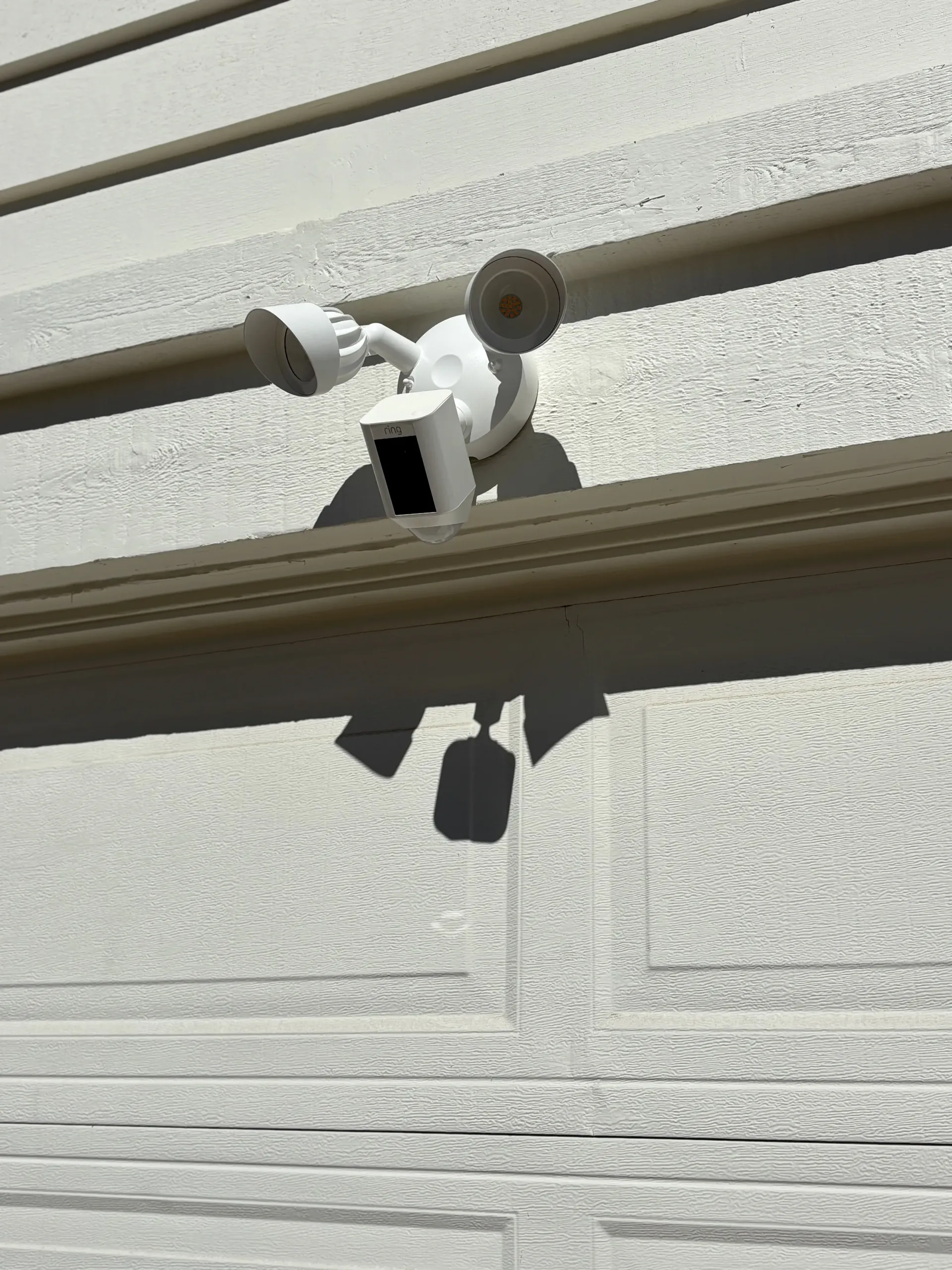 A white smart security camera with two floodlights mounted on a textured, light-colored garage wall.