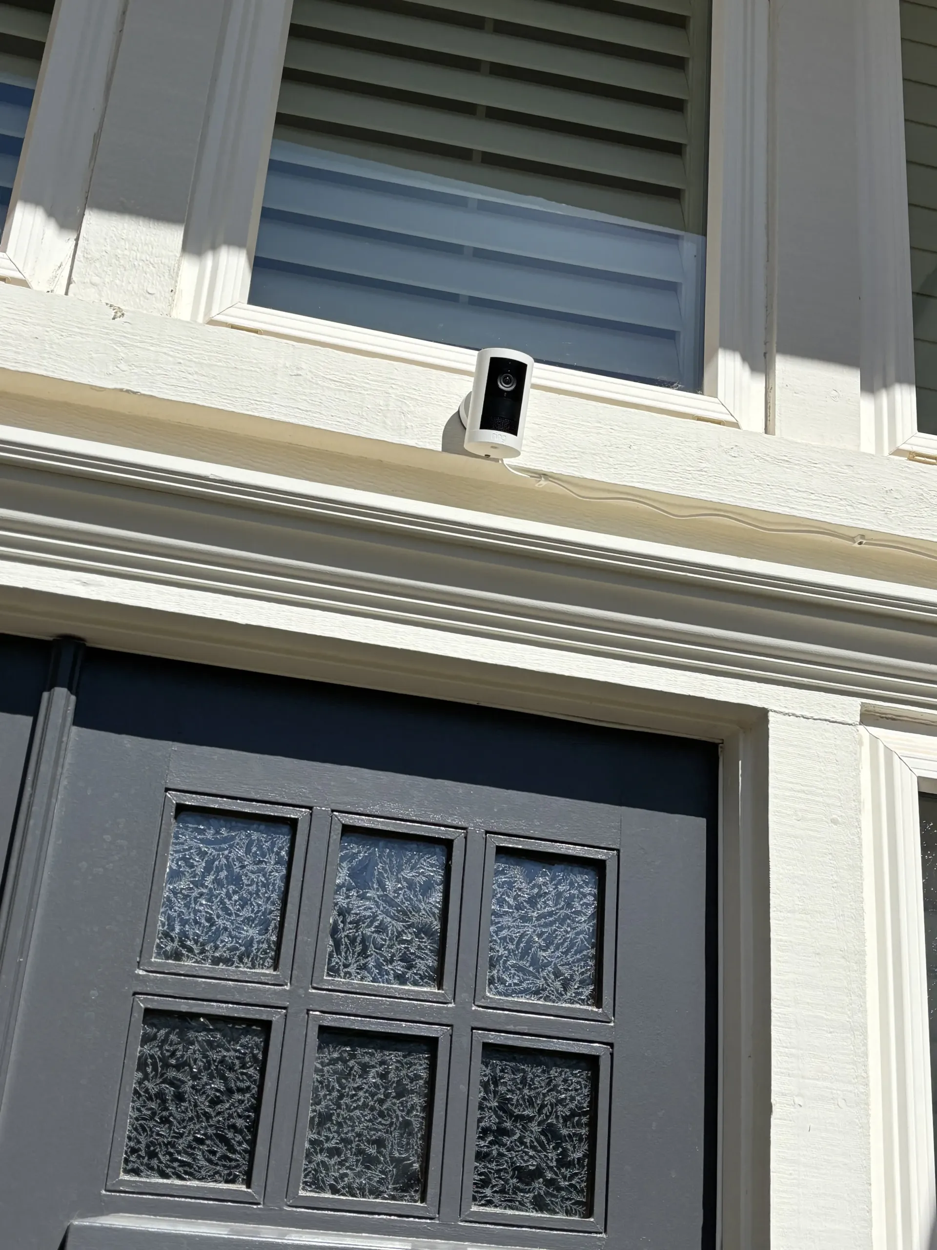 A white security camera mounted above a dark gray front door with a grid of six frosted glass window panes.