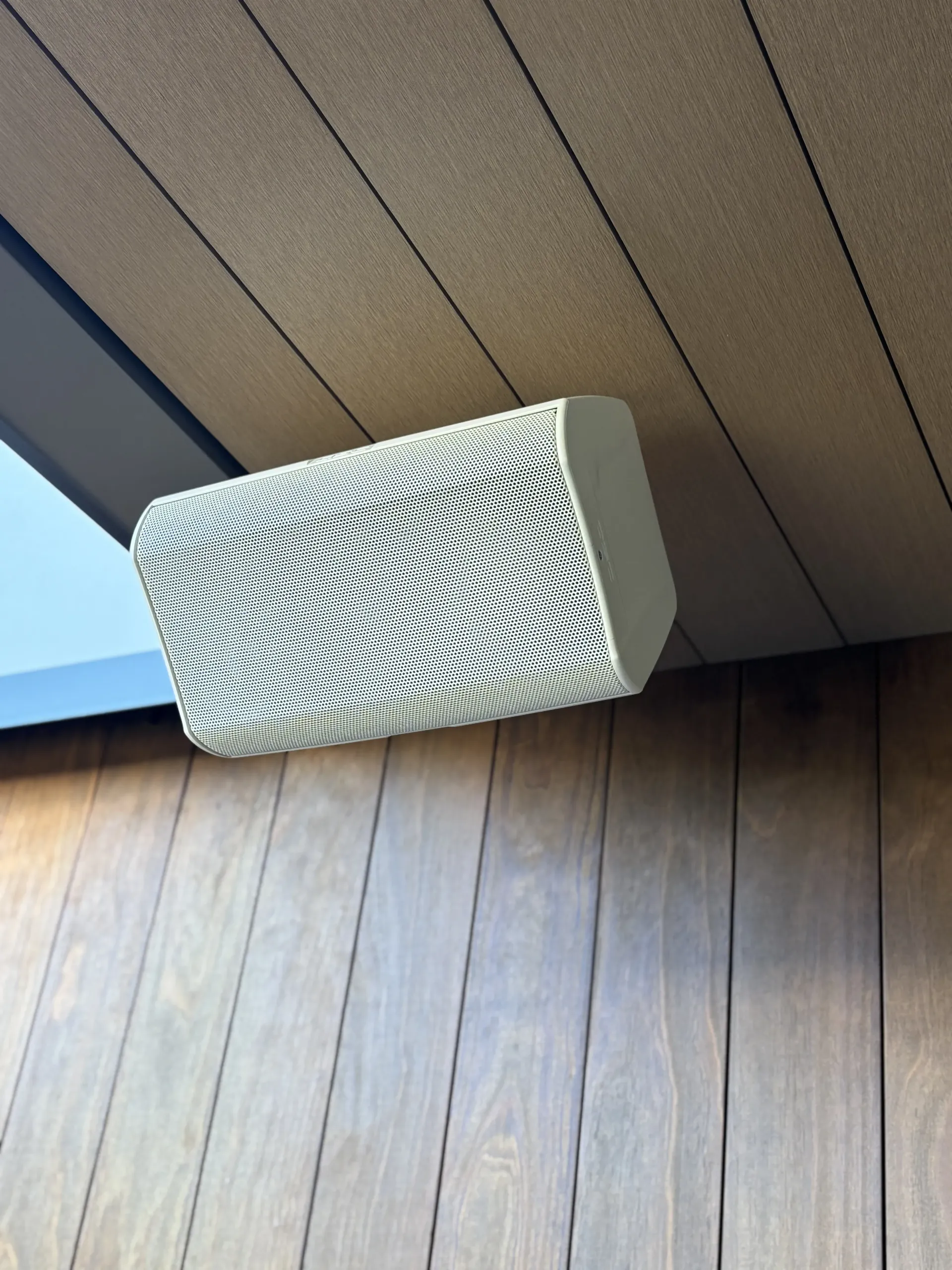 A white, textured outdoor speaker is mounted to a wooden-paneled ceiling near a glass window.