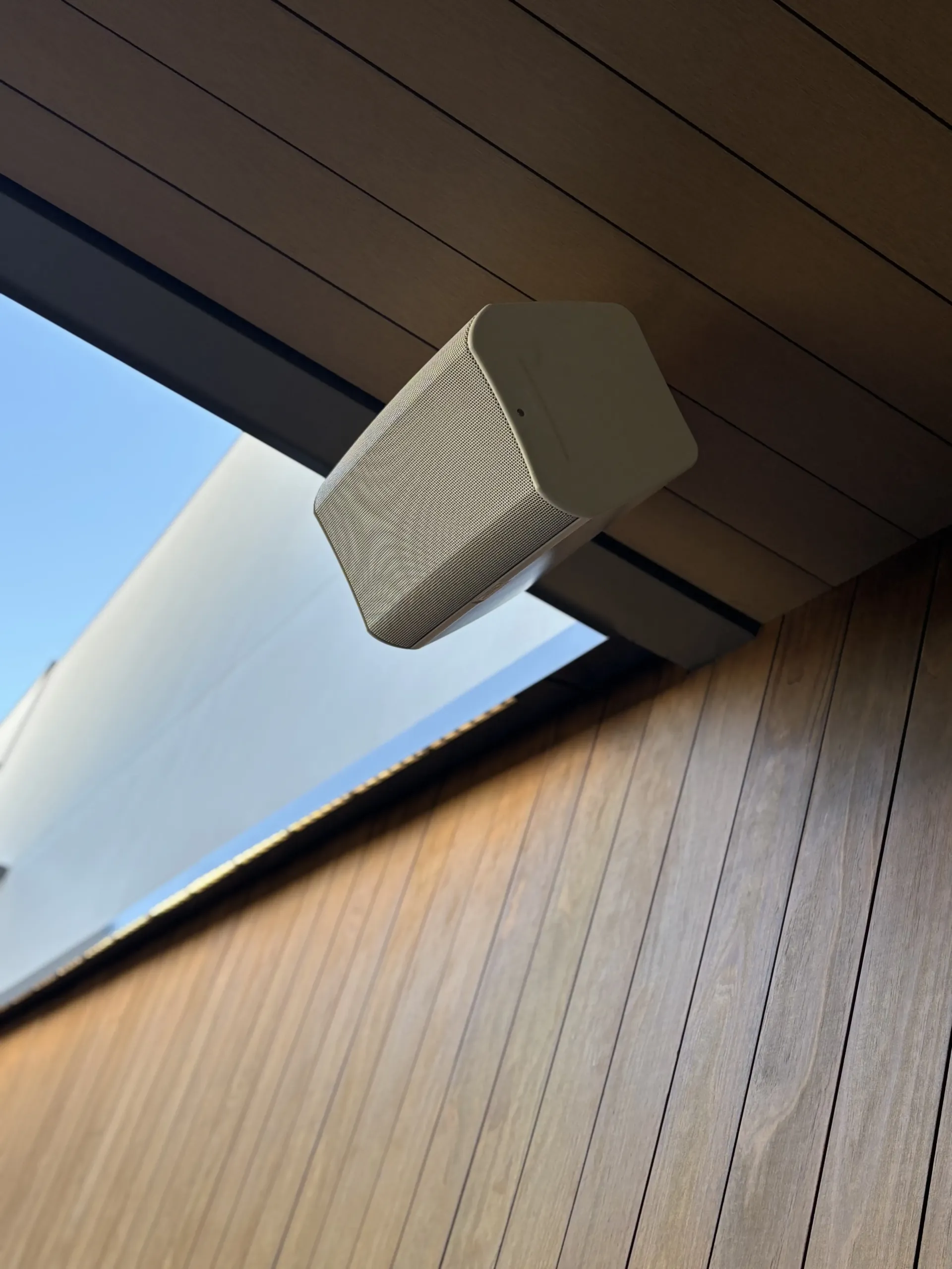 An off-white outdoor speaker mounted on the wooden ceiling of a porch, viewed from below against a blue sky.
