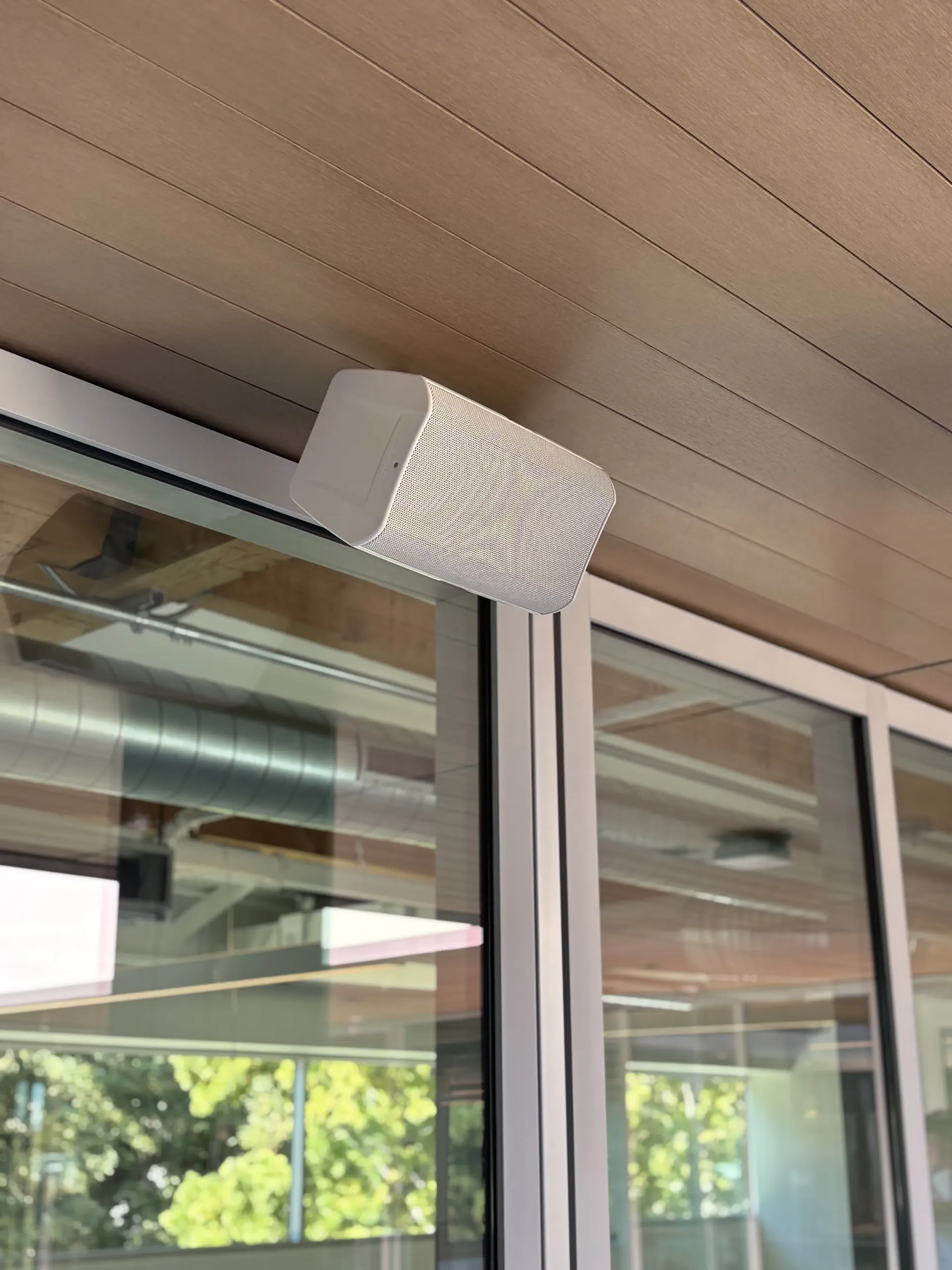 A white wall-mounted speaker positioned on a ceiling beam next to a glass window frame.