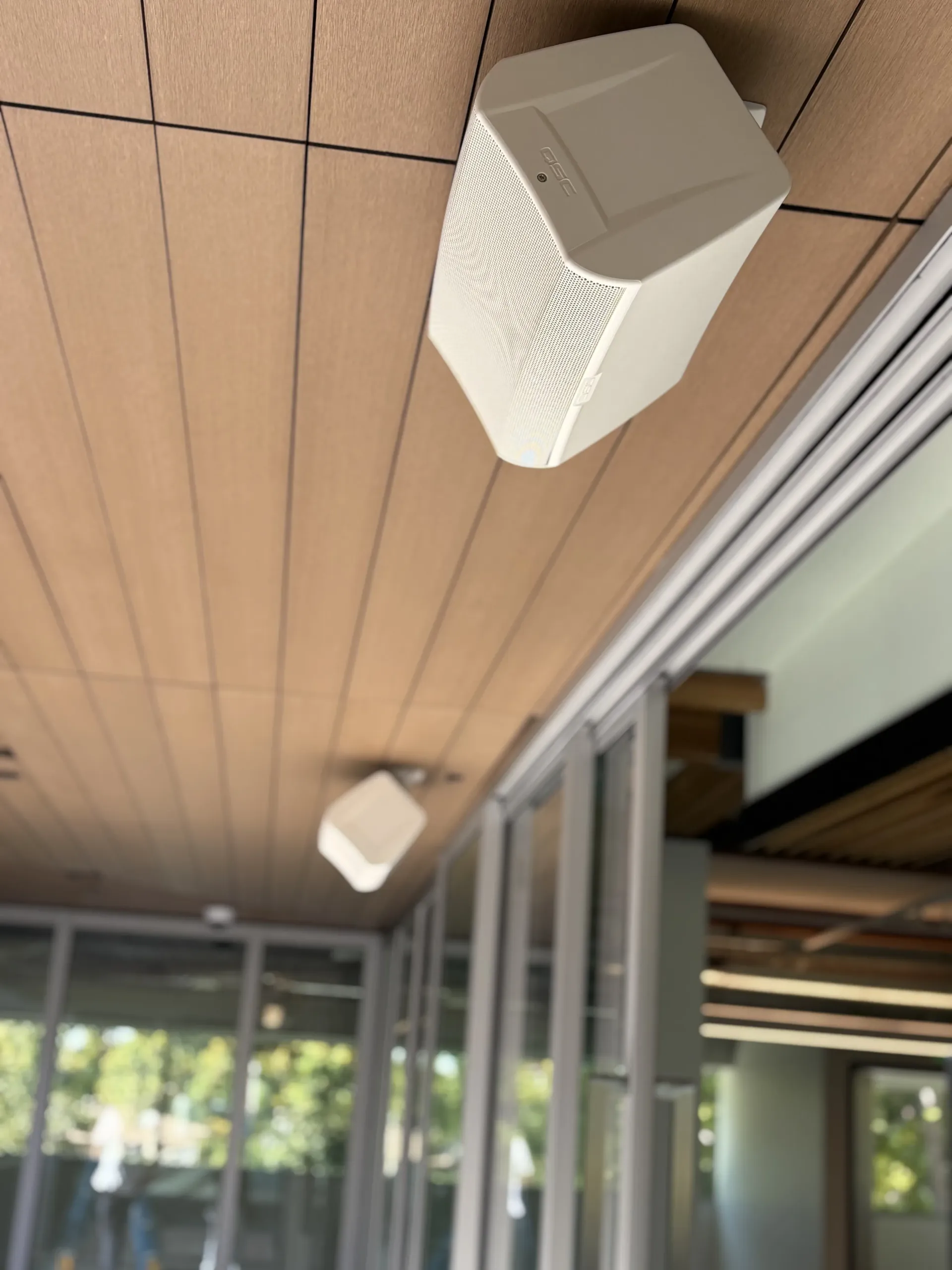 Two white, wall-mounted speakers attached to a wooden ceiling above a glass-walled structure.