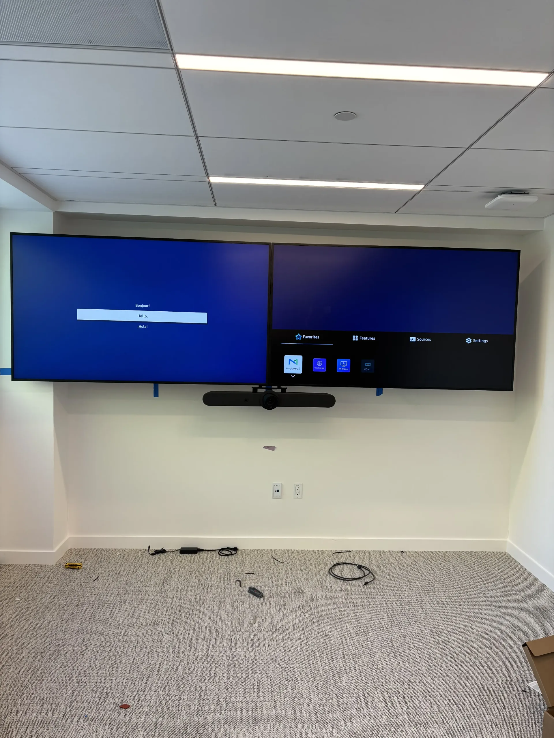 Two large, side-by-side monitors mounted on a wall above a soundbar in an office room, displaying a menu screen.