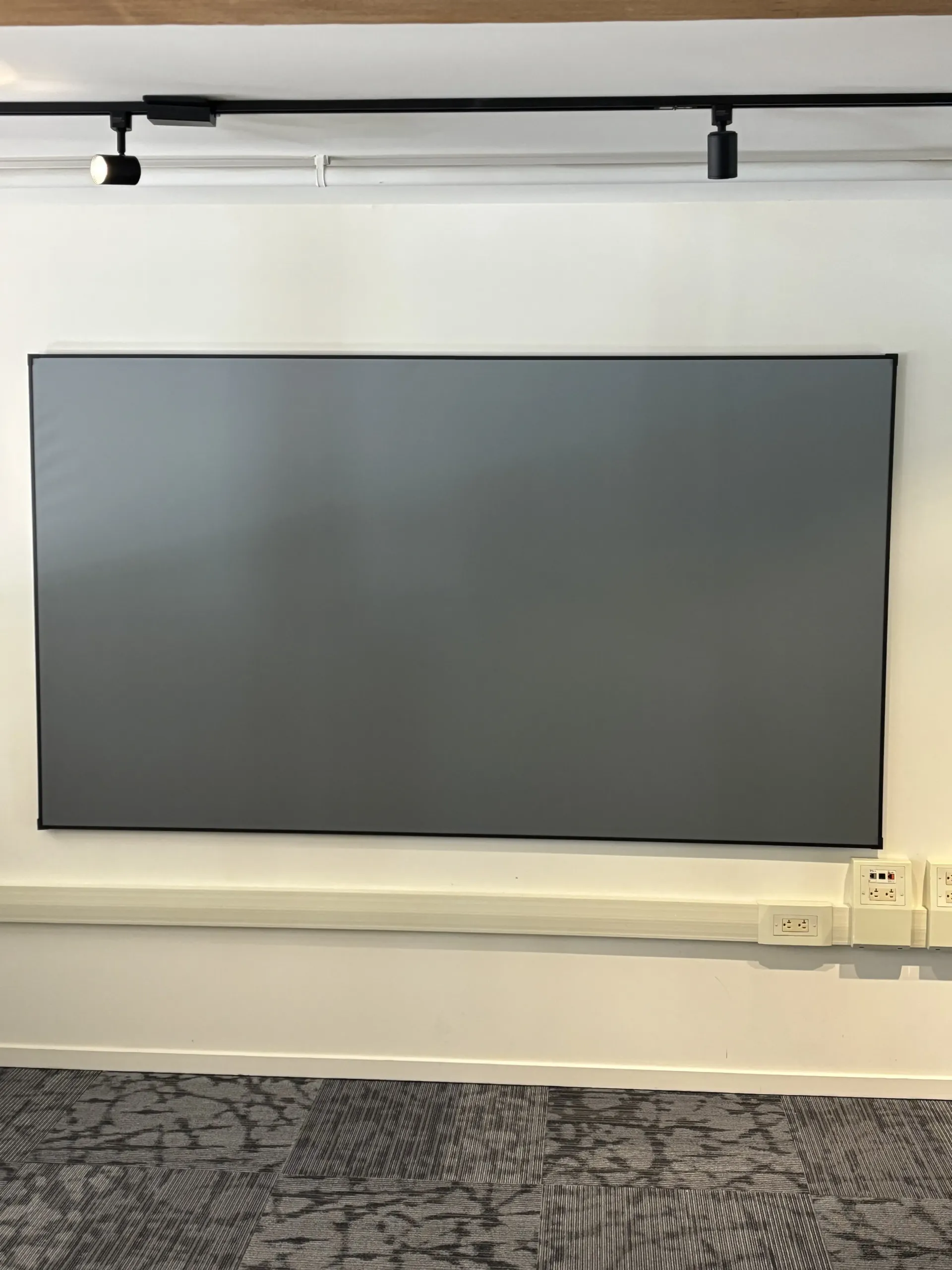 A gray, rectangular projection screen mounted on a white wall above electrical outlets, with a track light fixture above it.