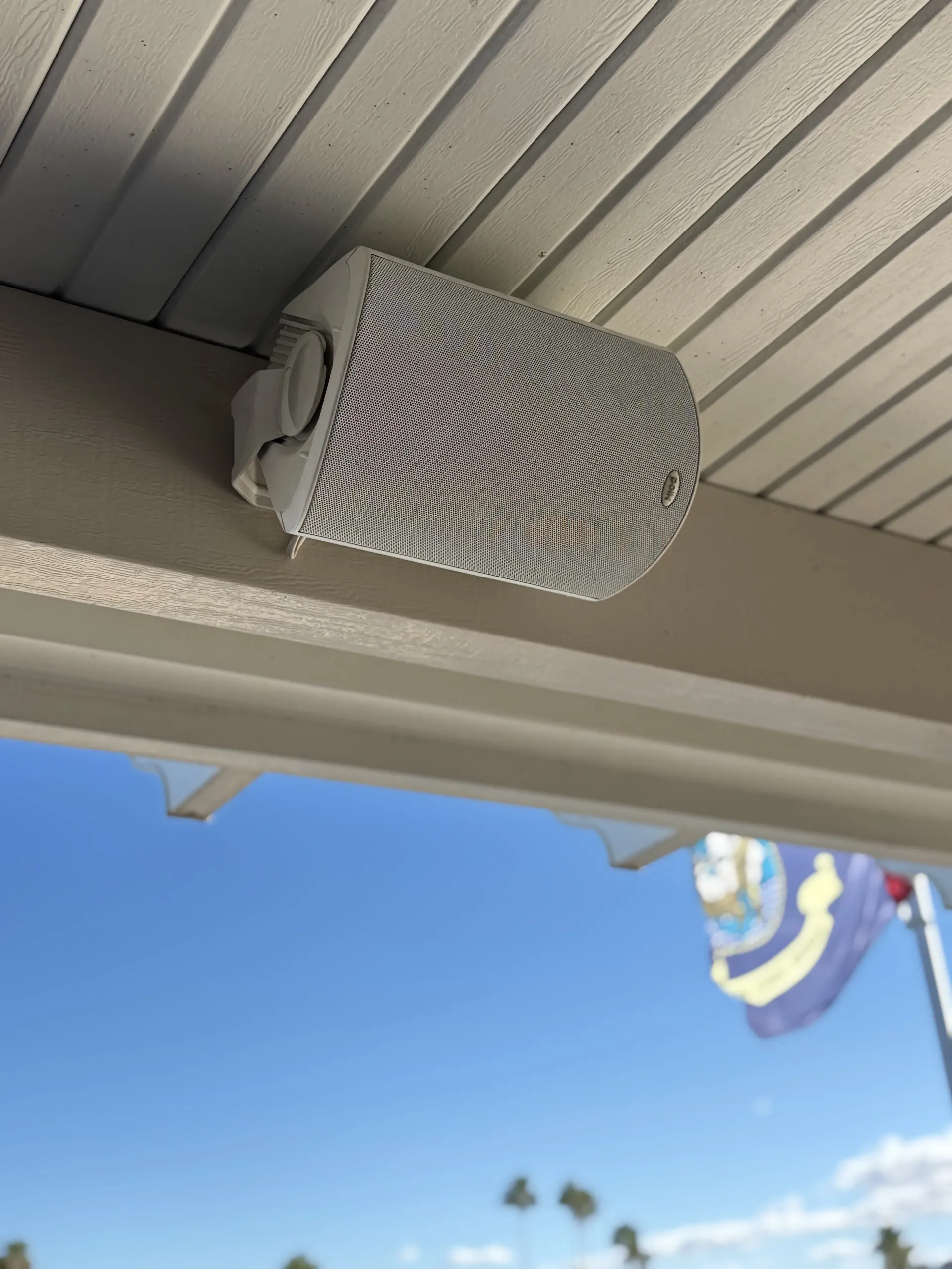A white outdoor speaker mounted to the ceiling and wooden beam of a covered patio, with a blue sky visible in the distance.