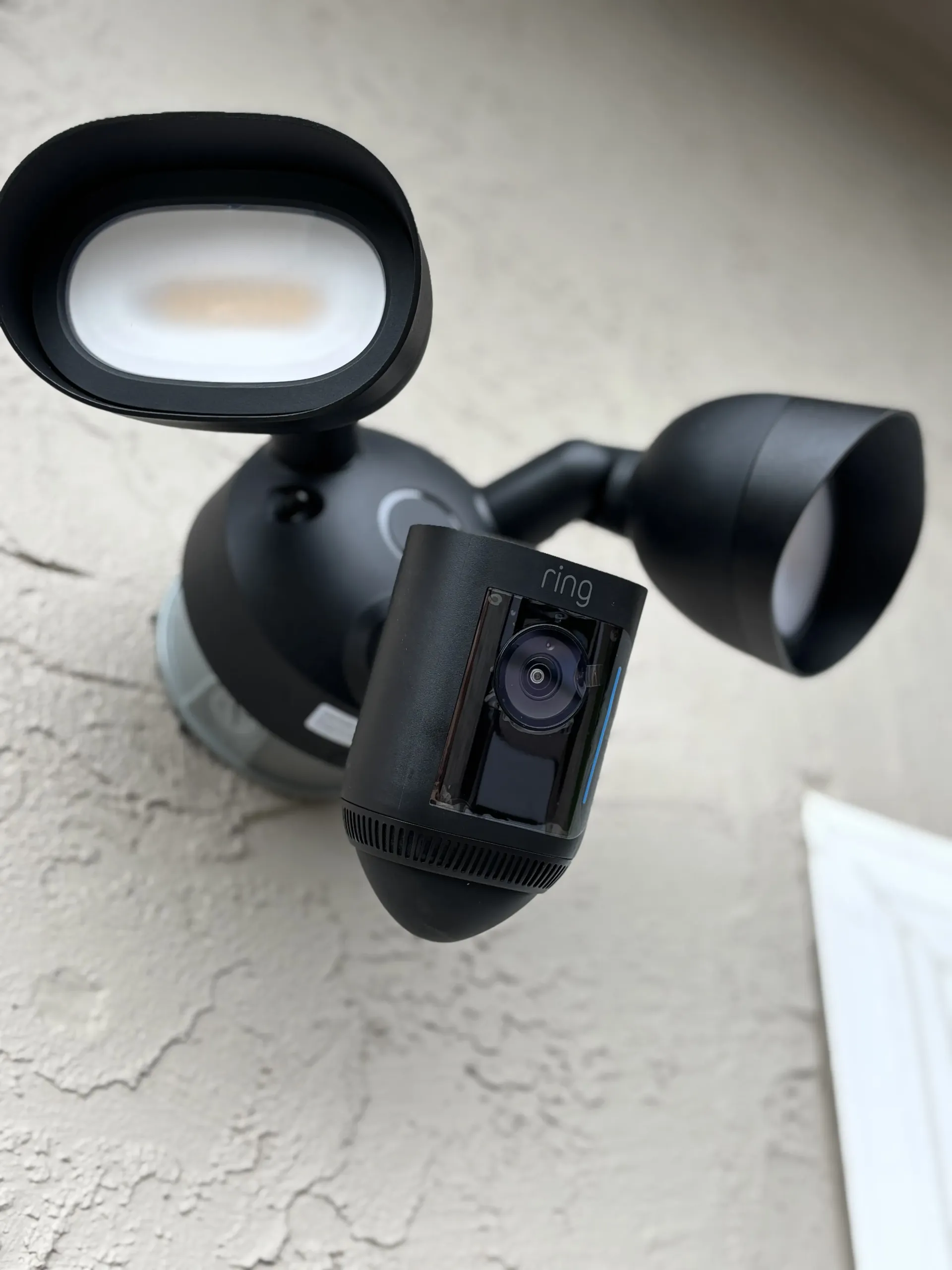A black Ring floodlight security camera mounted on a textured, light-colored wall.
