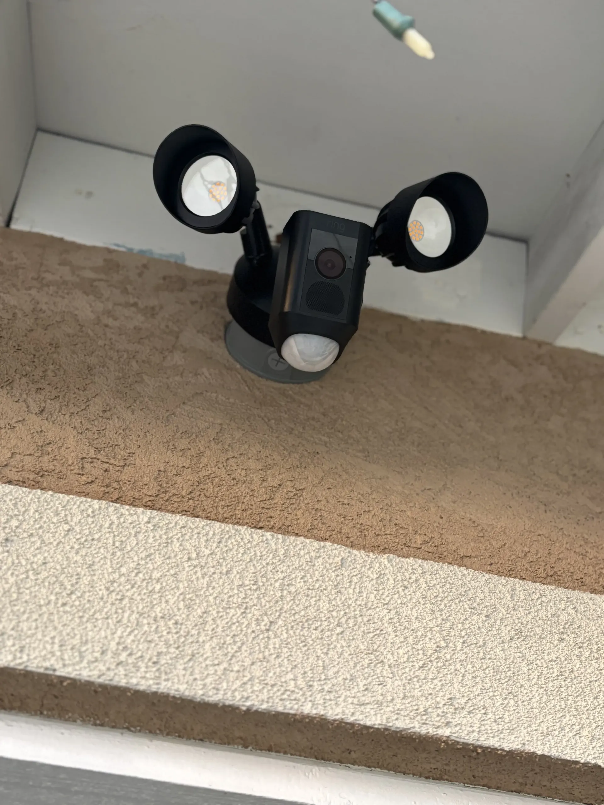 A black, dual-head outdoor motion-sensor floodlight with an integrated camera mounted on a textured tan wall.