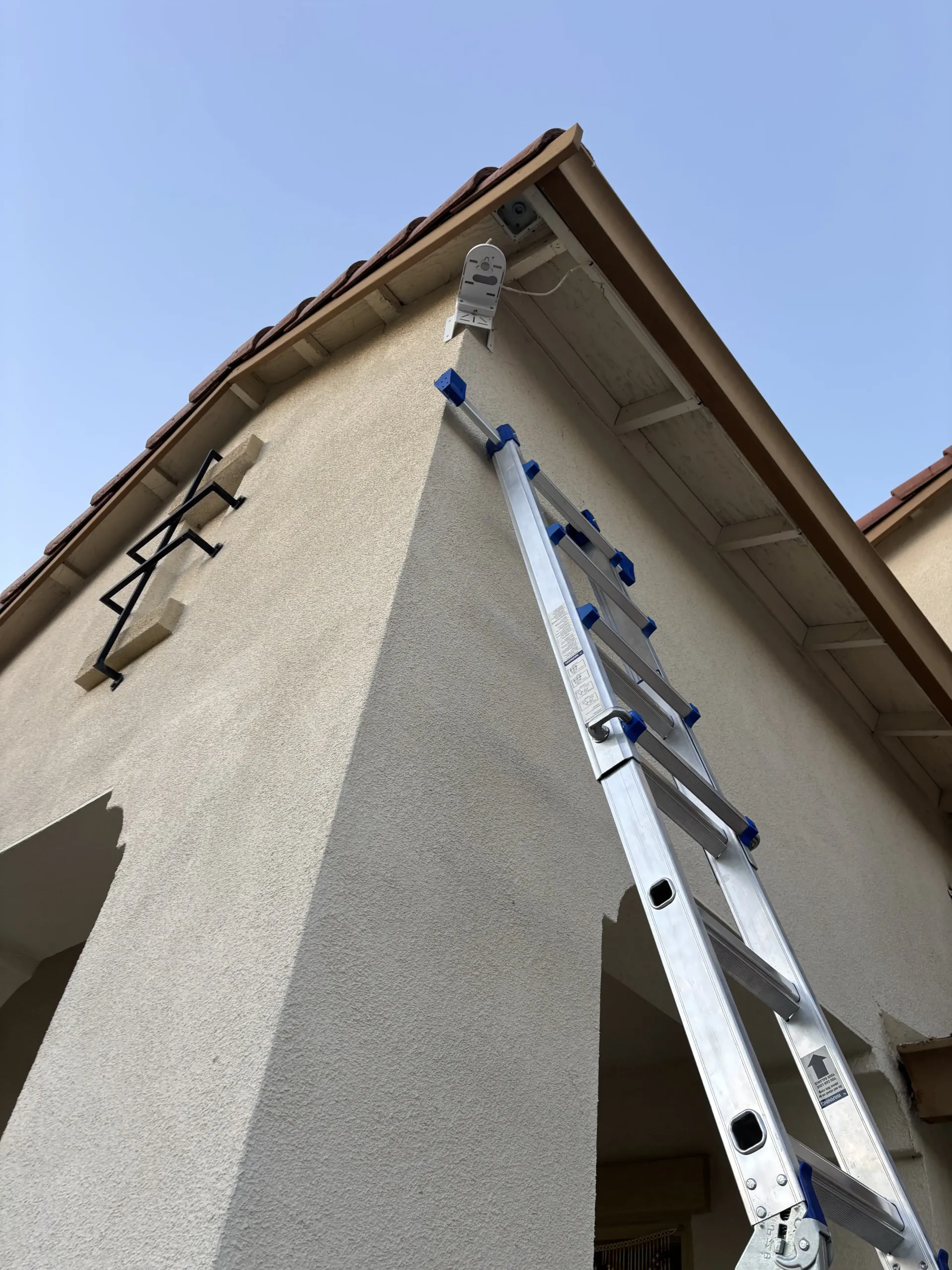 An aluminum extension ladder leans against the corner of a tan stucco building under a clear blue sky.