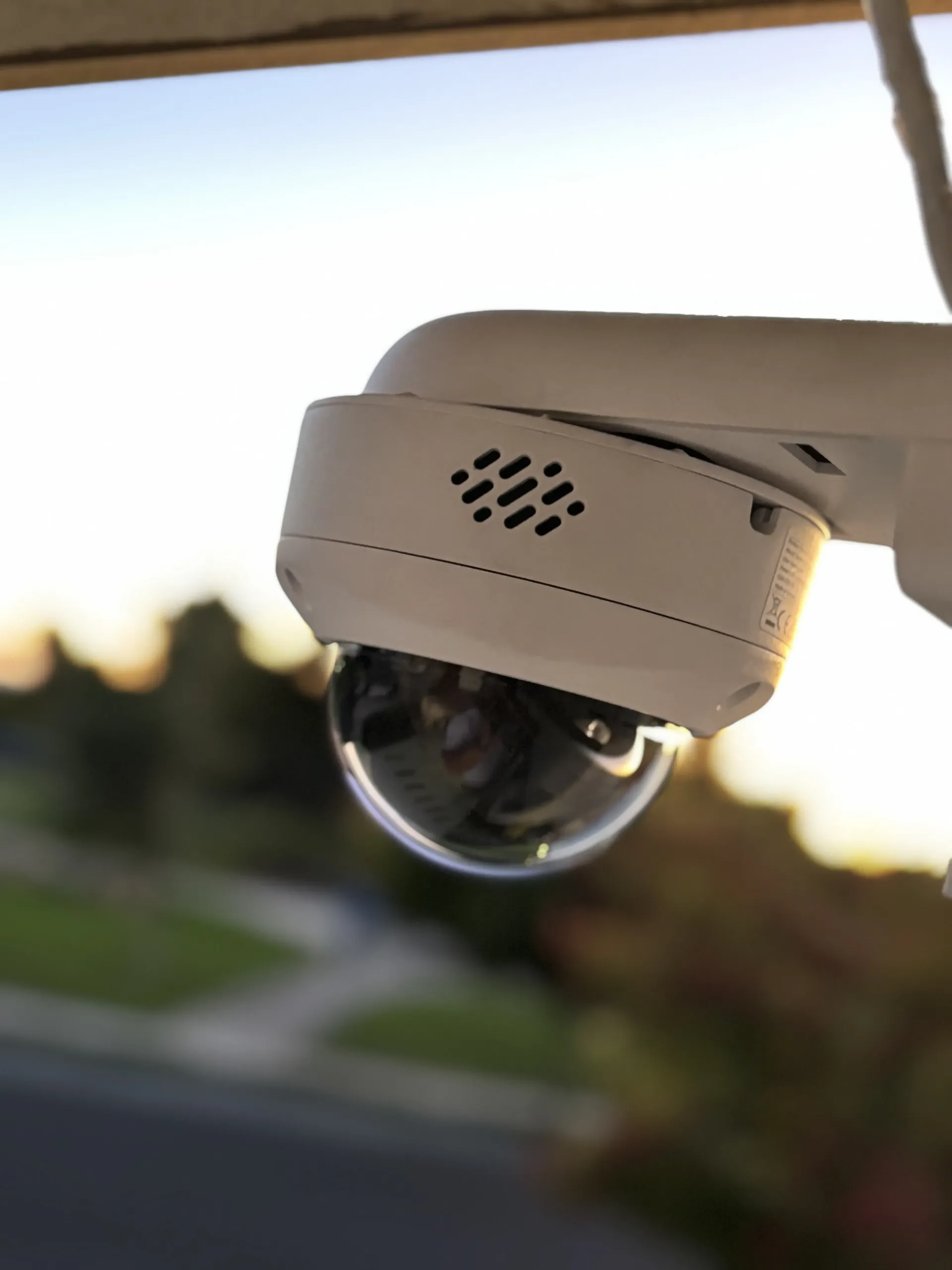 A white, dome-style security camera mounted under an eave, pointed toward an outdoor residential street at sunset.