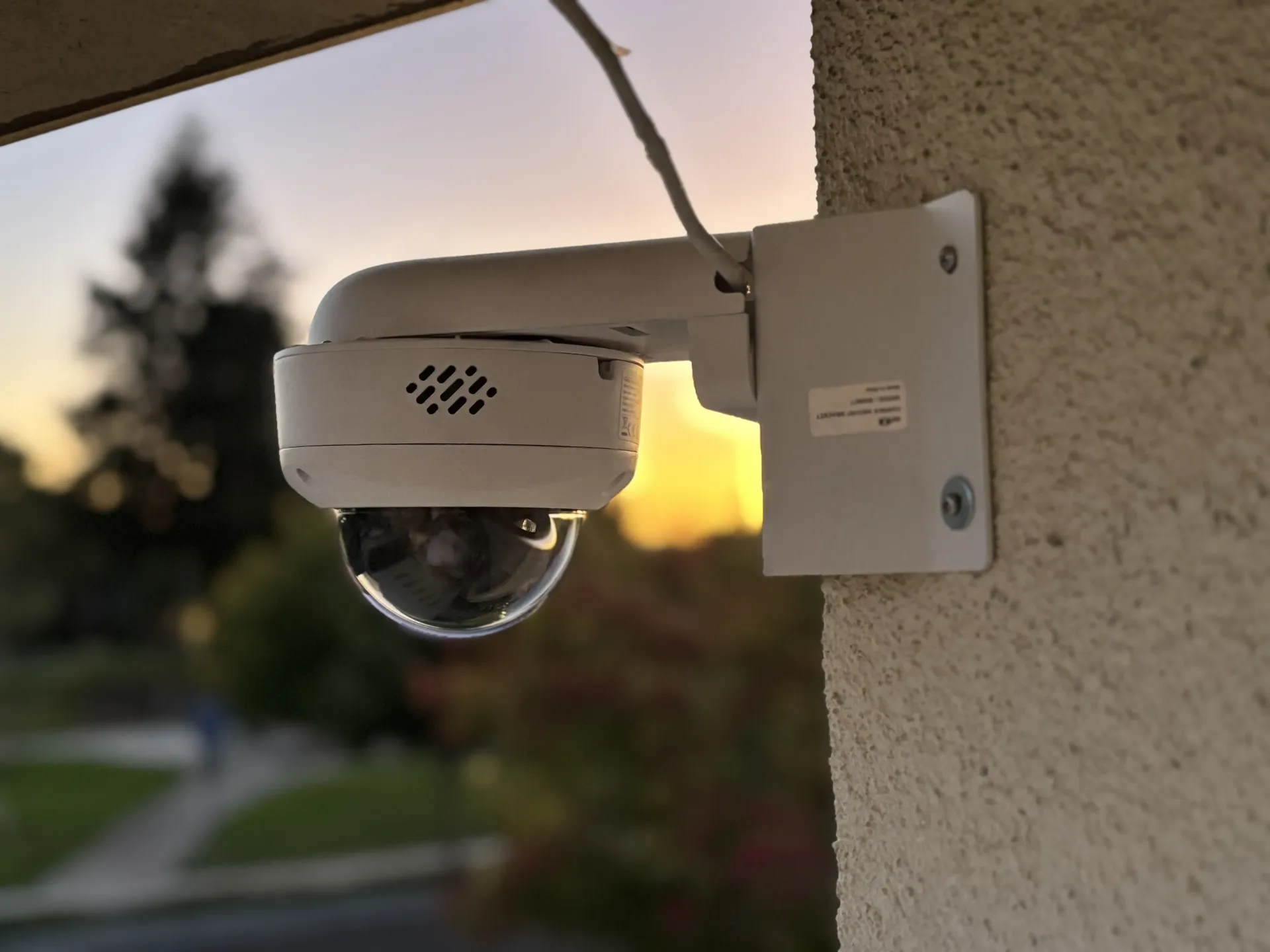 A white dome security camera mounted to an exterior wall during a sunset.