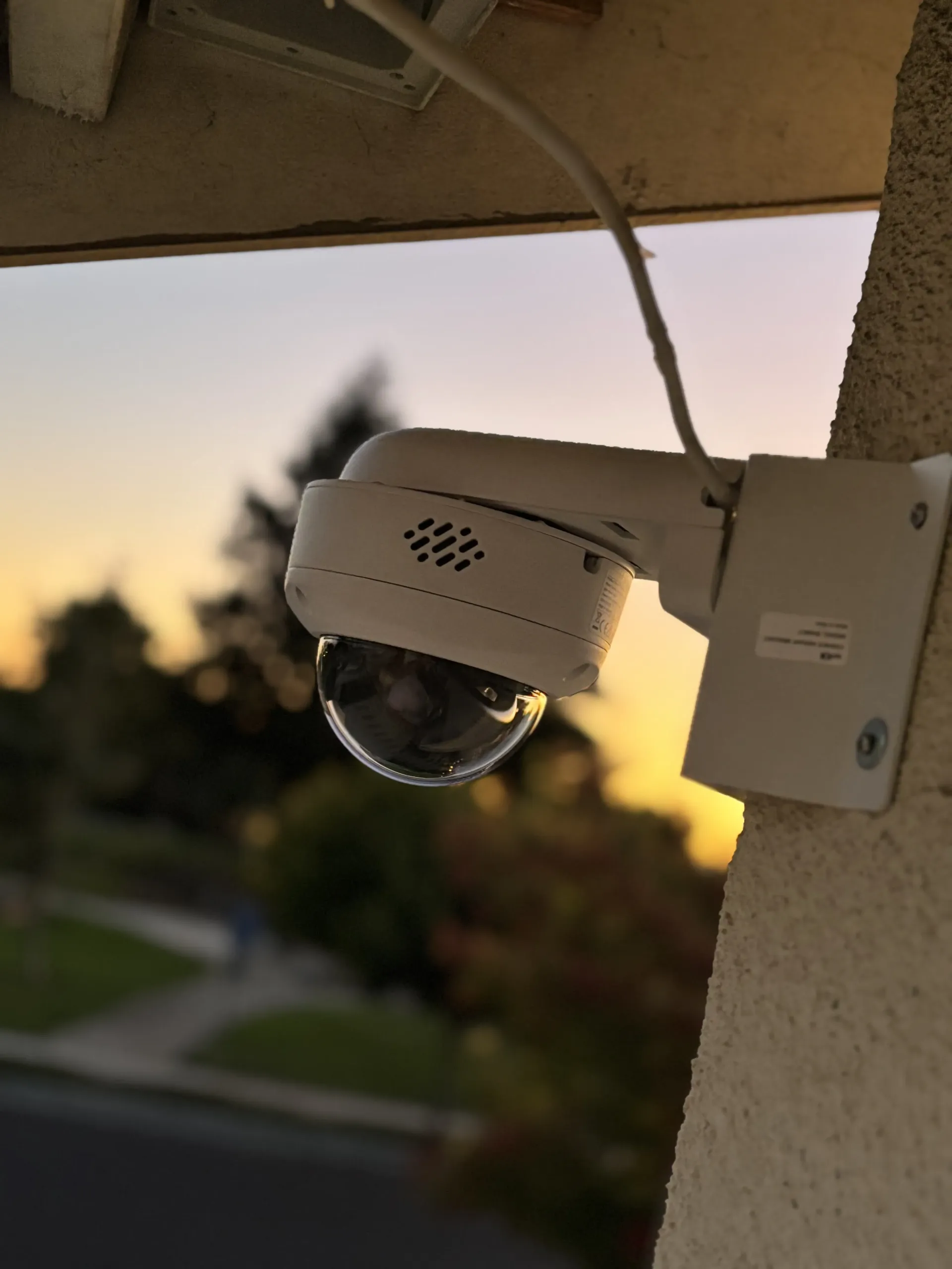 A white dome security camera mounted on a textured wall against a sunset sky.