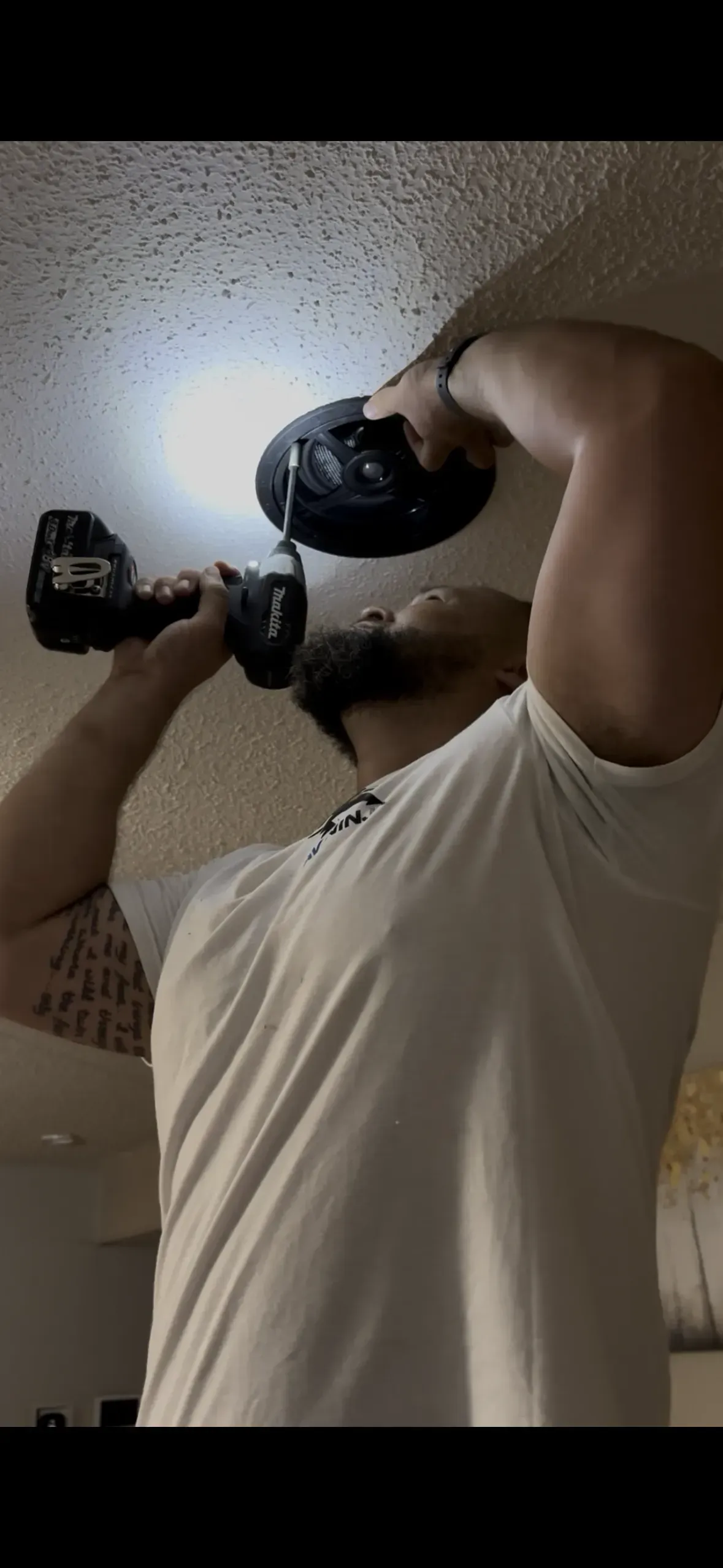 A person in a white t-shirt uses a cordless drill to install a circular light fixture onto a textured ceiling.