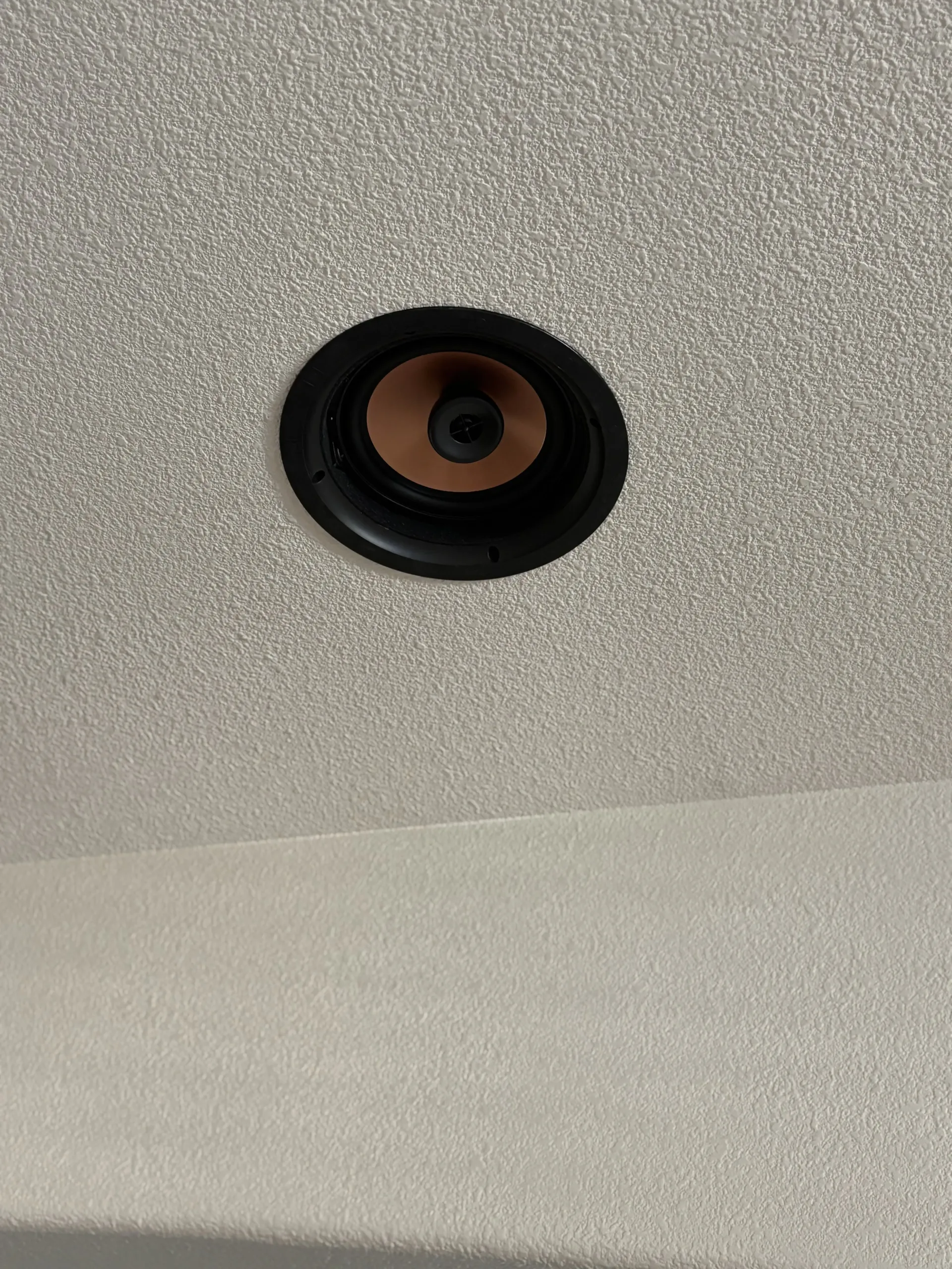 A circular, black recessed speaker mounted in a textured, light gray ceiling.