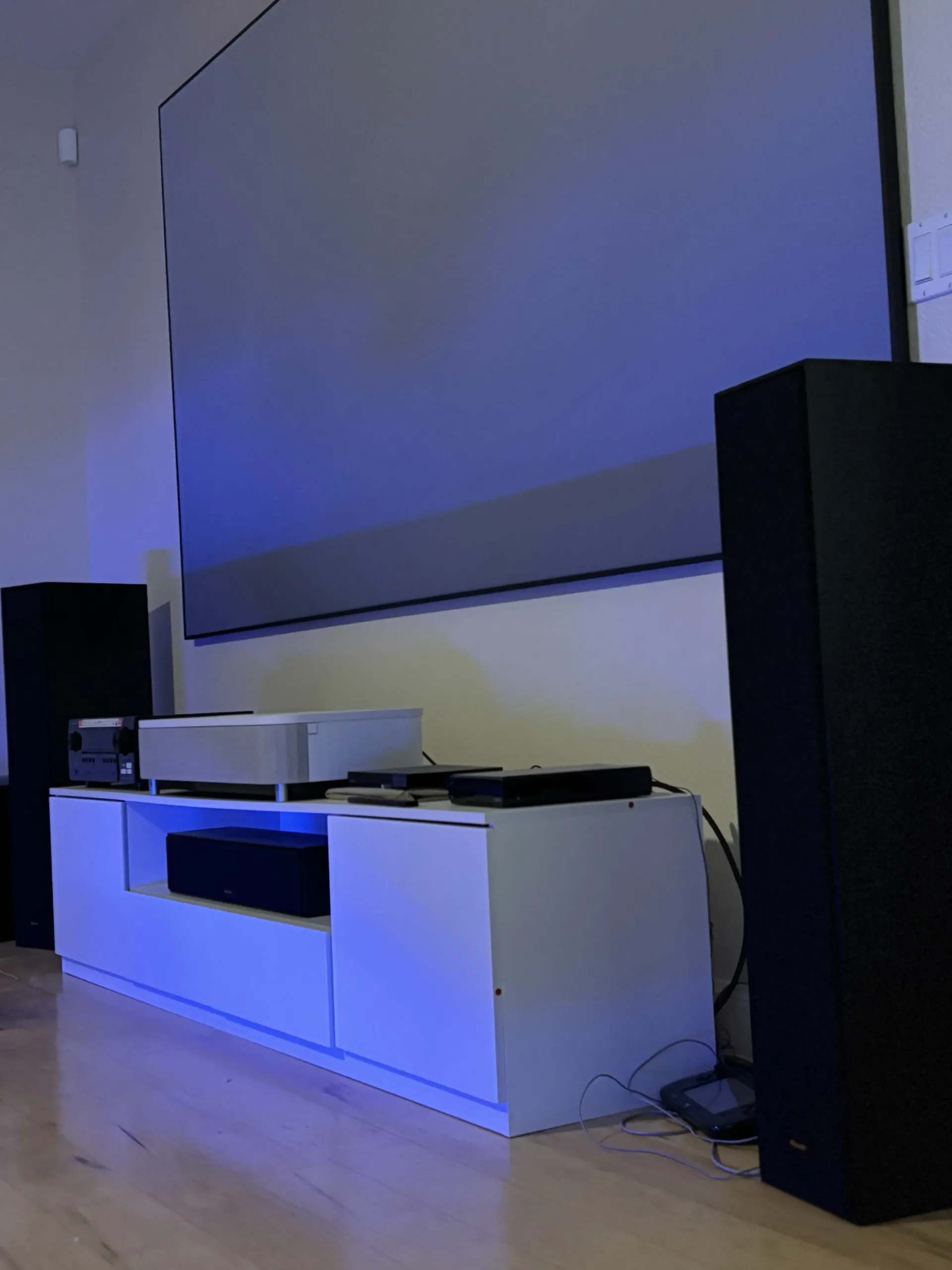 A home theater setup with a large projection screen, two black tower speakers, and a white media console with electronics.