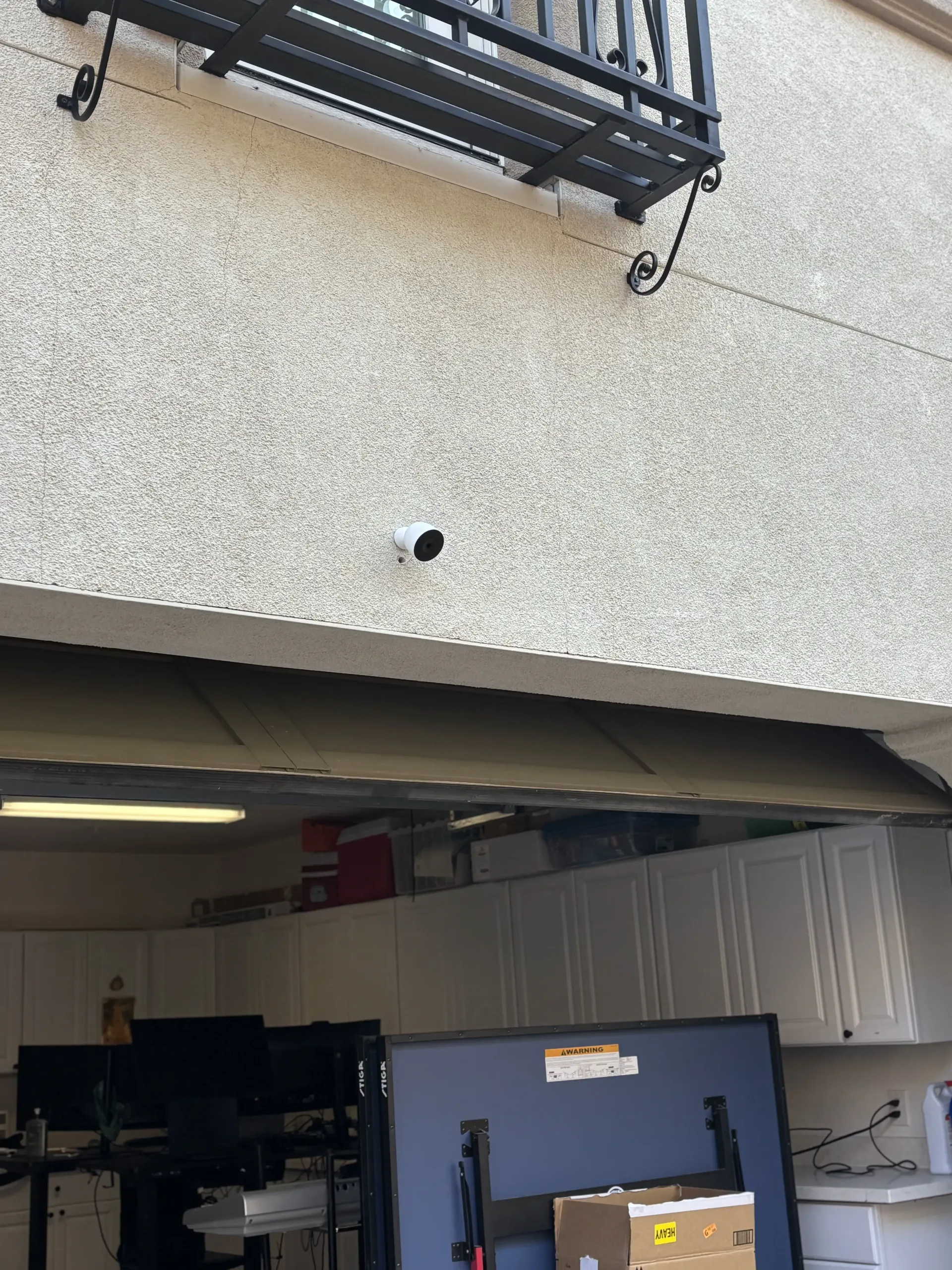 A white security camera mounted on the stucco exterior wall above an open garage containing storage cabinets and equipment.