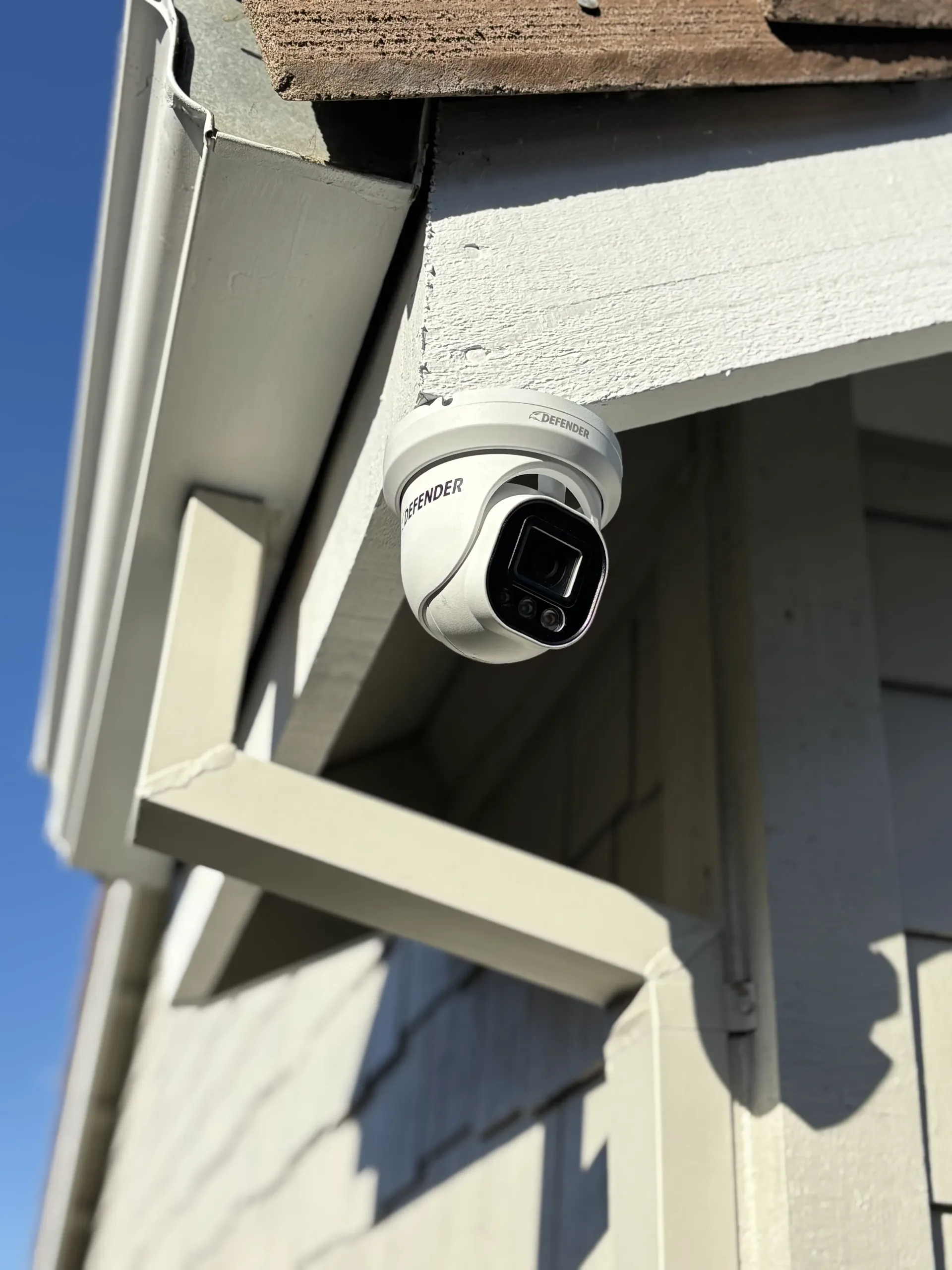 A white dome-style security camera mounted under the white wooden eave of a building against a clear blue sky.