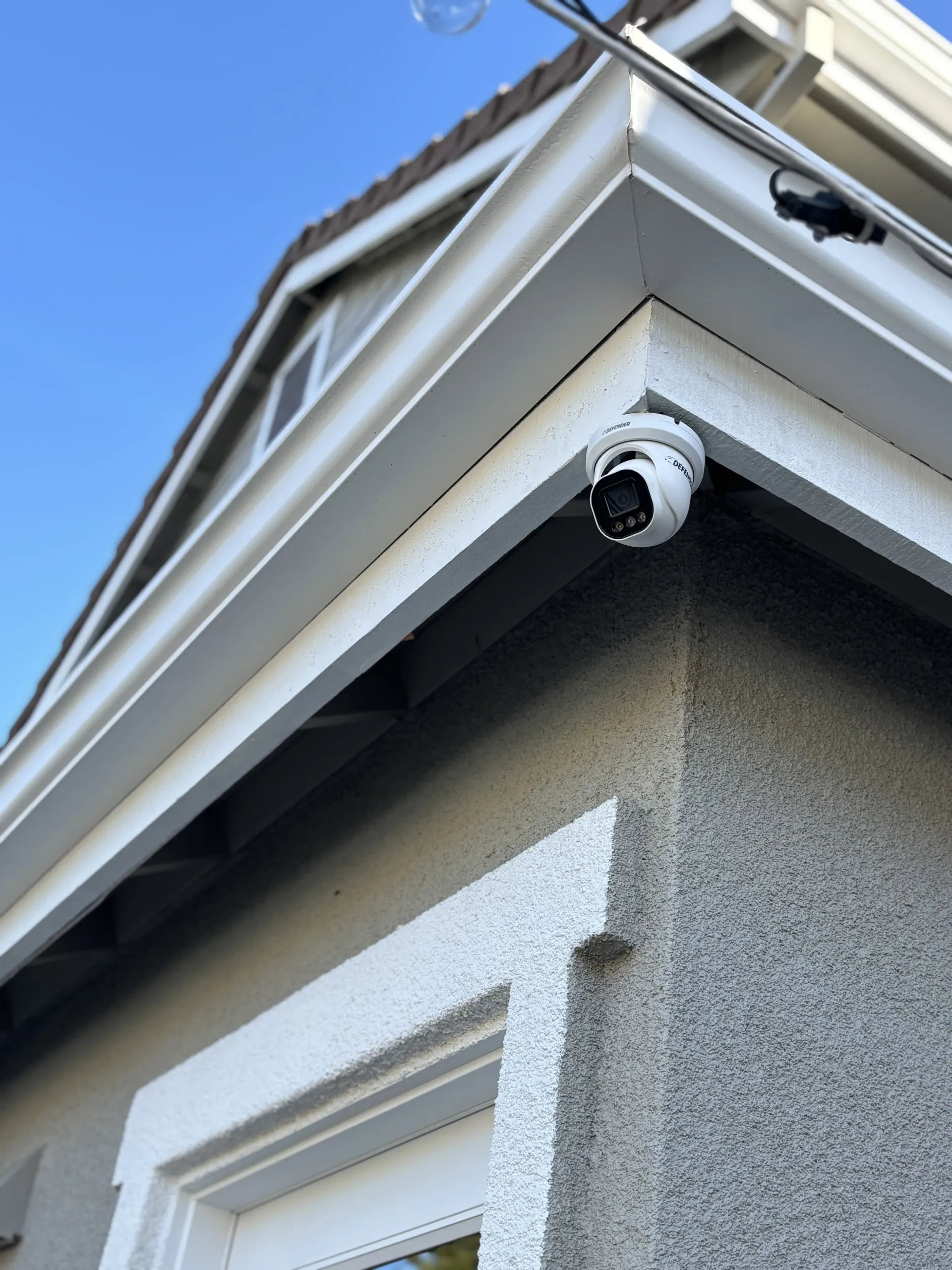 A white security dome camera mounted on the corner of a grey stucco house under a bright blue sky.