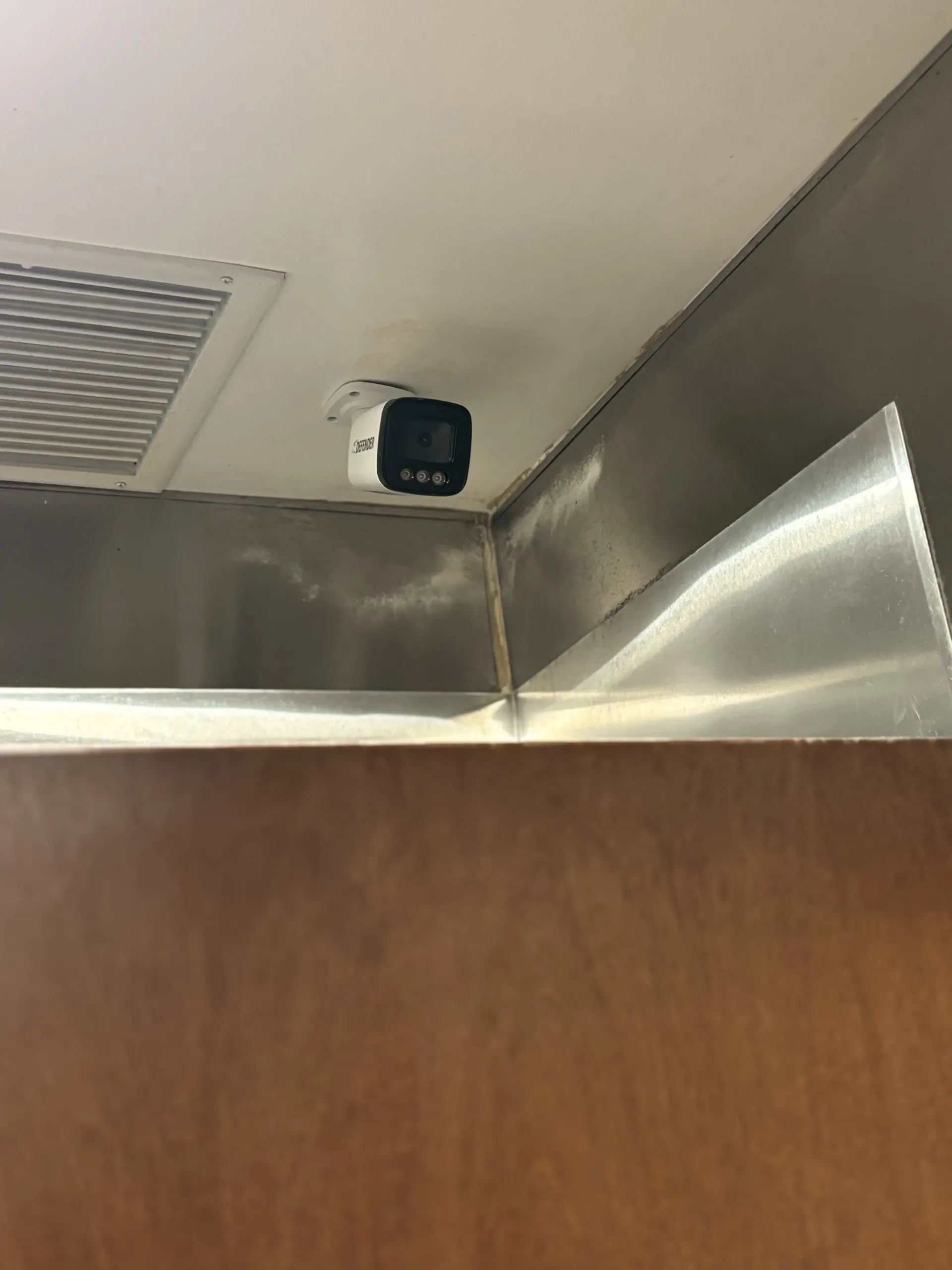 A security camera mounted on a ceiling near a metallic vent above a wooden door frame.
