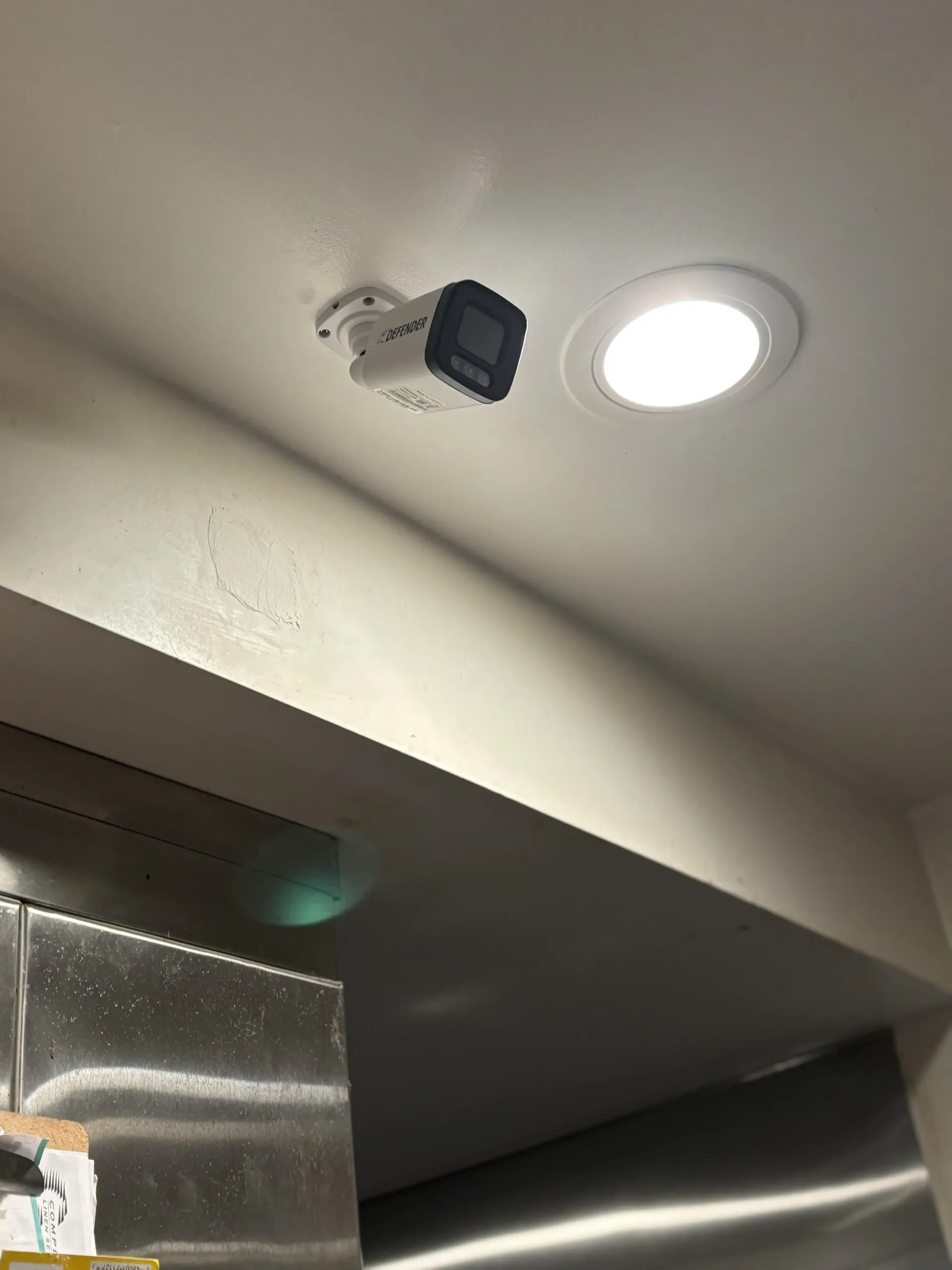 A white security camera mounted on a white ceiling next to a recessed circular light fixture.