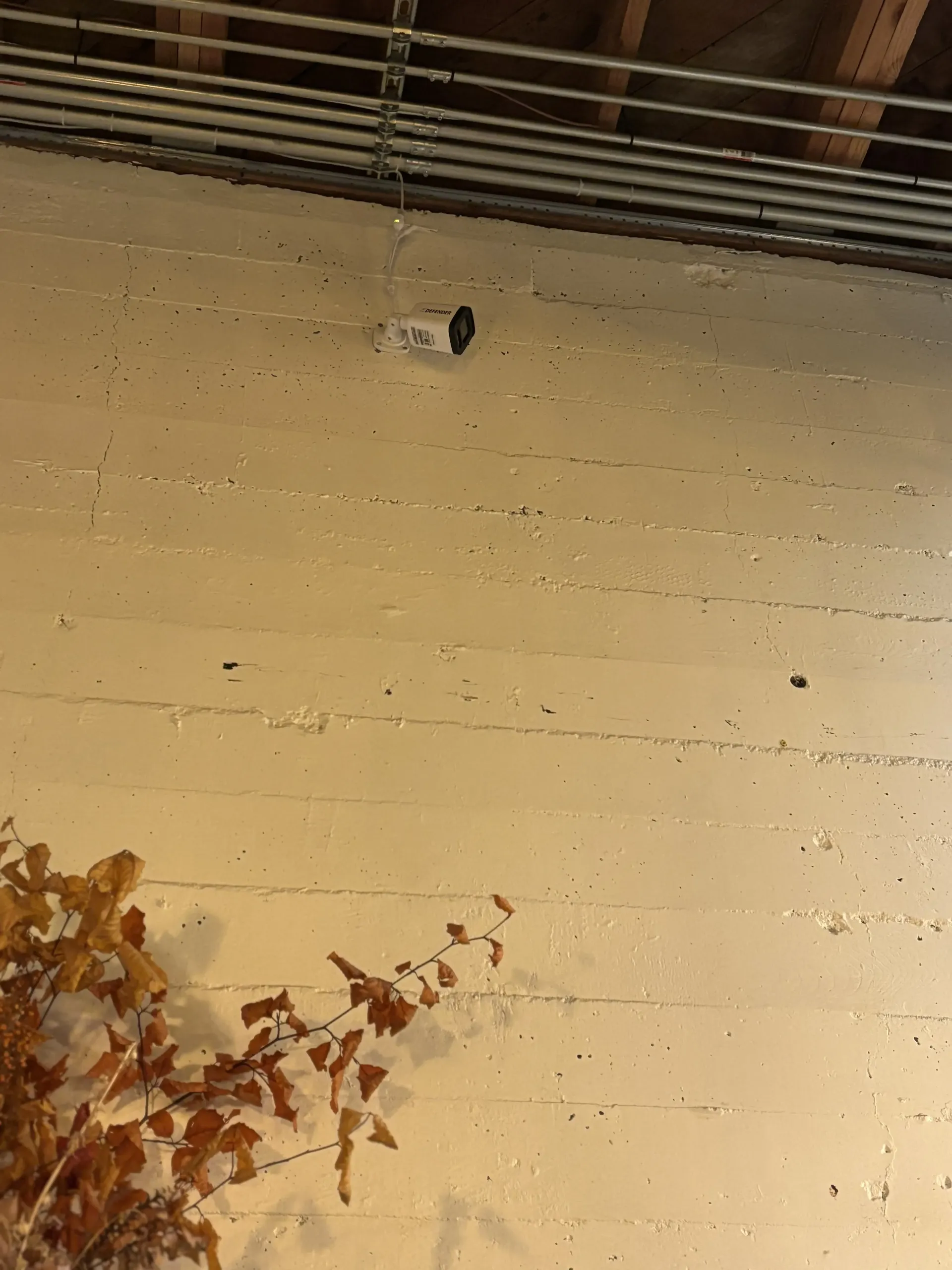 A security camera mounted on a beige, textured wall below exposed metal pipes and wooden rafters.