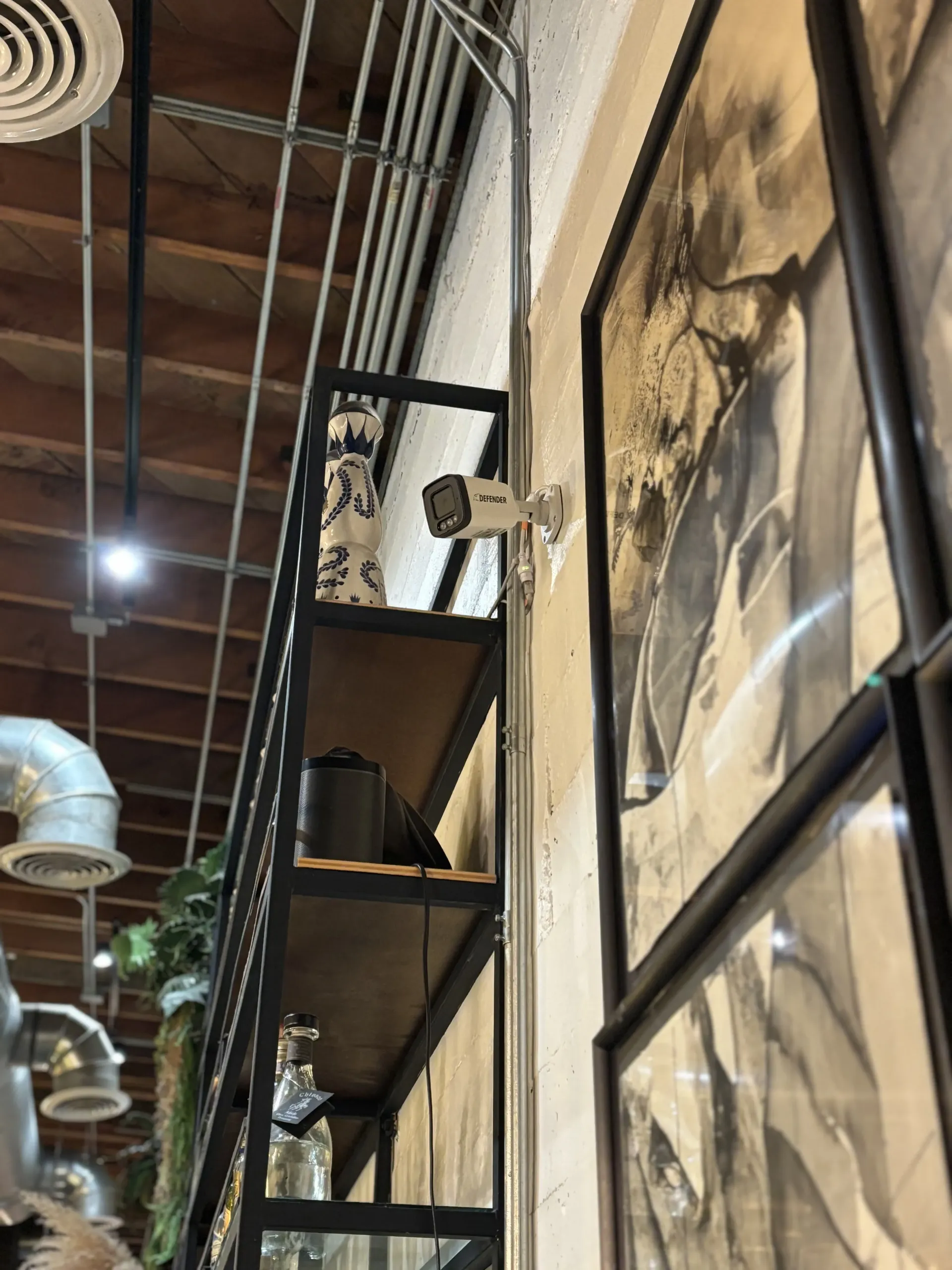 A security camera mounted on a wall next to a black metal shelf and framed wall art in a room with an industrial ceiling.