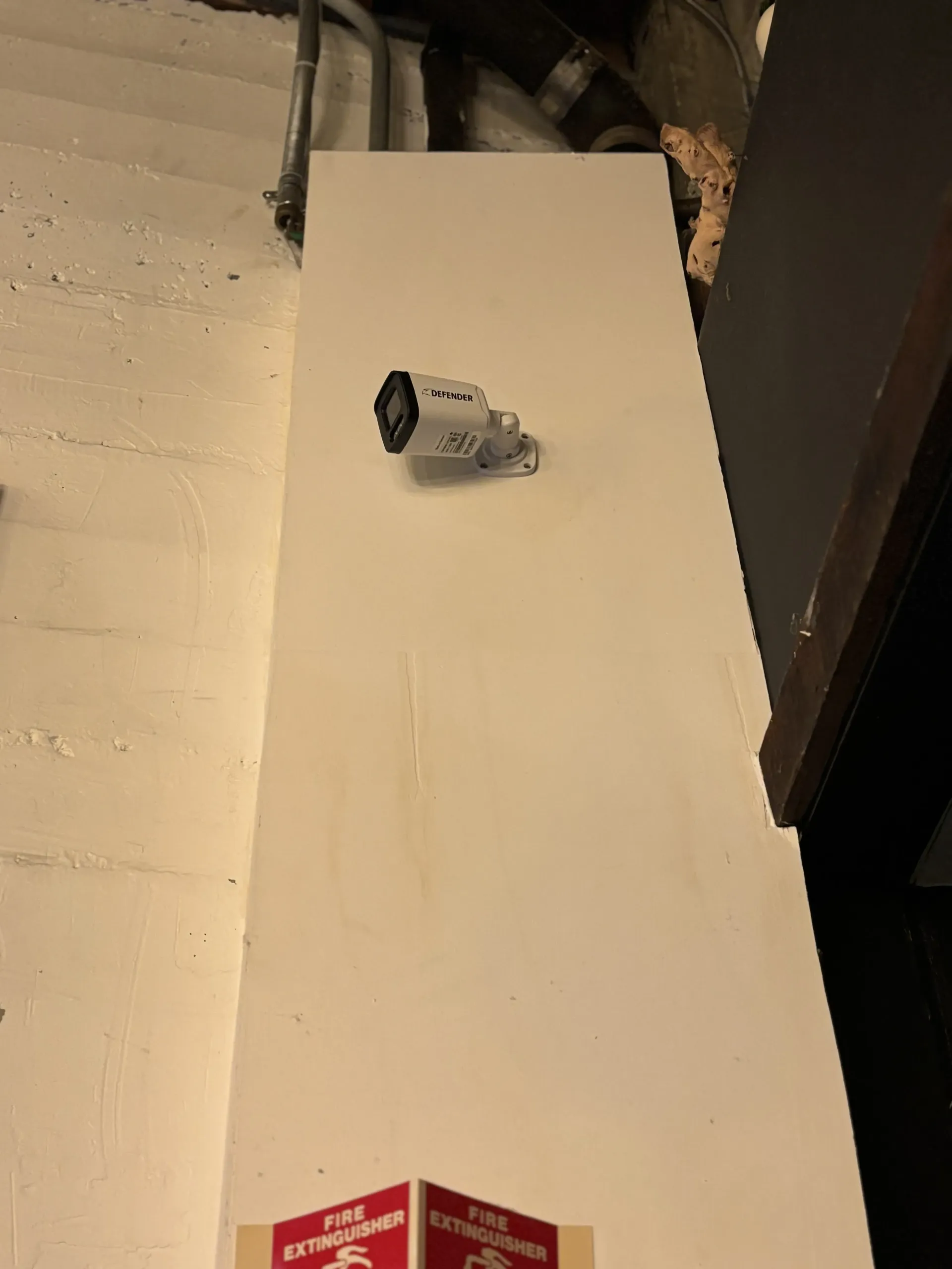 A security camera mounted on a white wall above a fire extinguisher sign in an indoor setting.