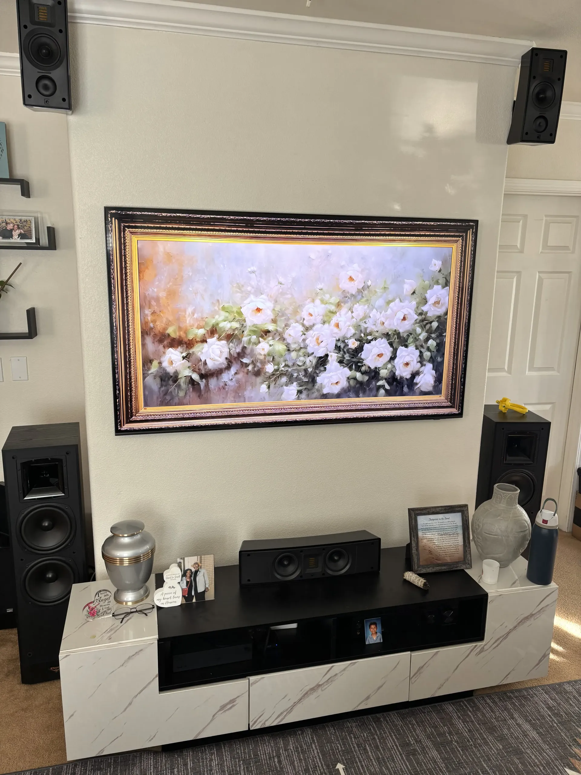 A living room TV wall with a large floral artwork, floor speakers, wall-mounted speakers, and a marble-finish console.