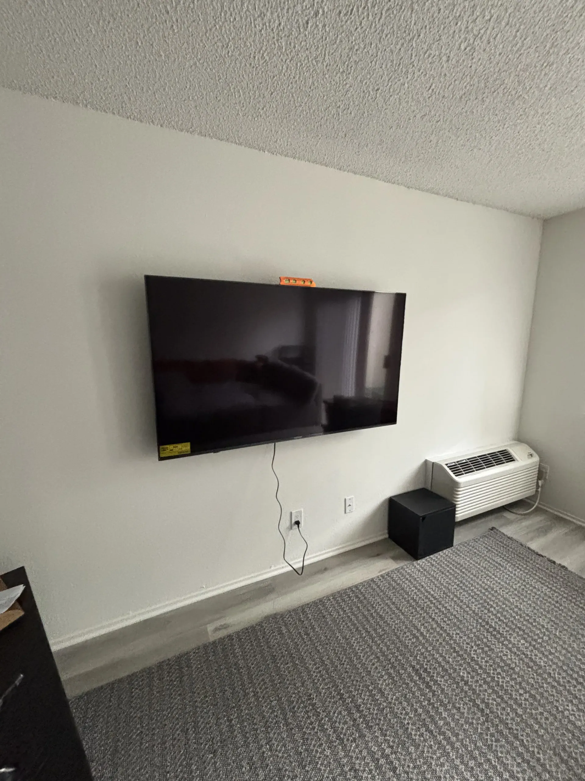 A wall-mounted television hangs in a room with light grey walls, a textured ceiling, a grey rug, and a small space heater.
