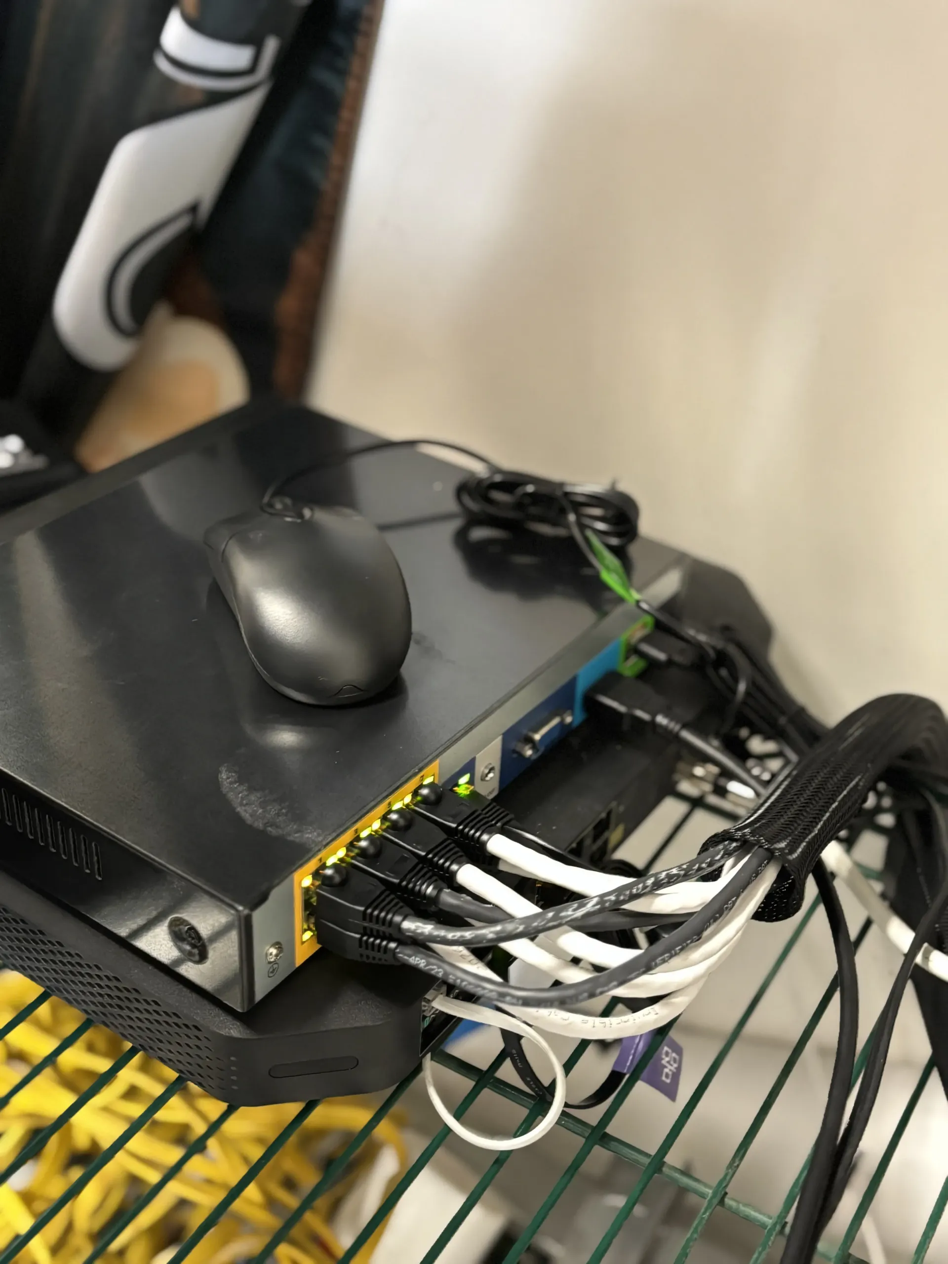 A black network switch with multiple Ethernet cables plugged into its front sits on a wire shelf with a mouse on top.