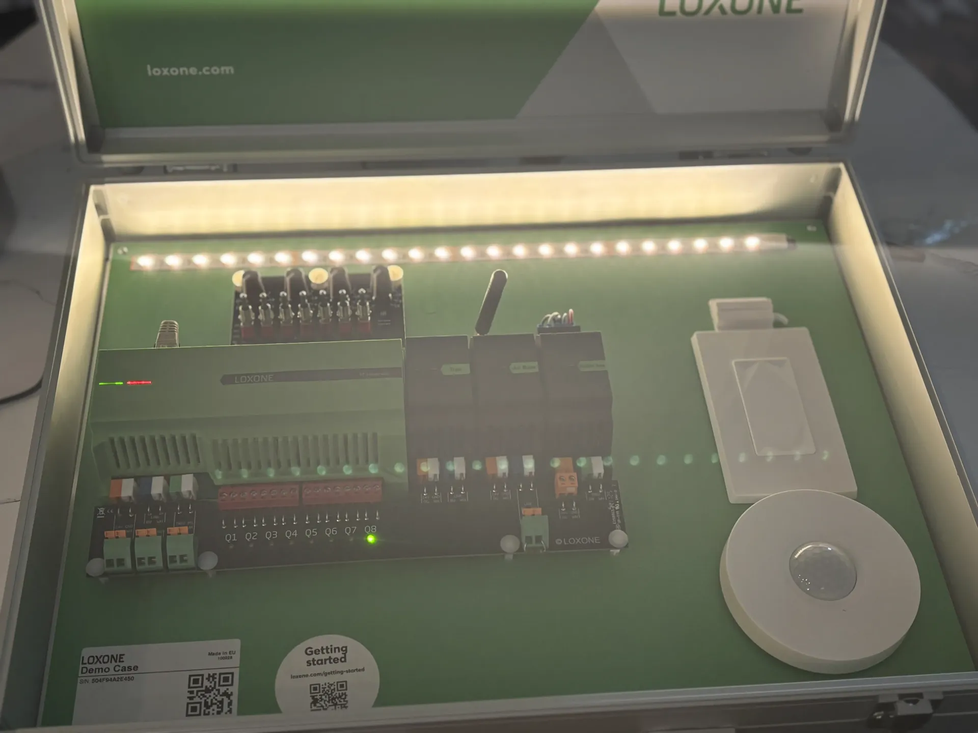 A Loxone smart home demo kit in a metallic case, featuring a controller, light strip, and motion sensor on a green board.