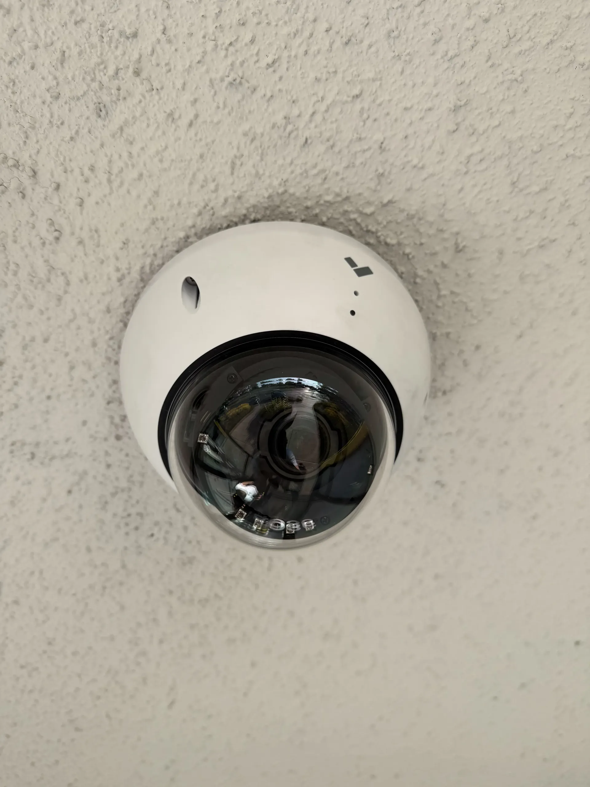 A small white dome security camera mounted on a textured off-white ceiling.