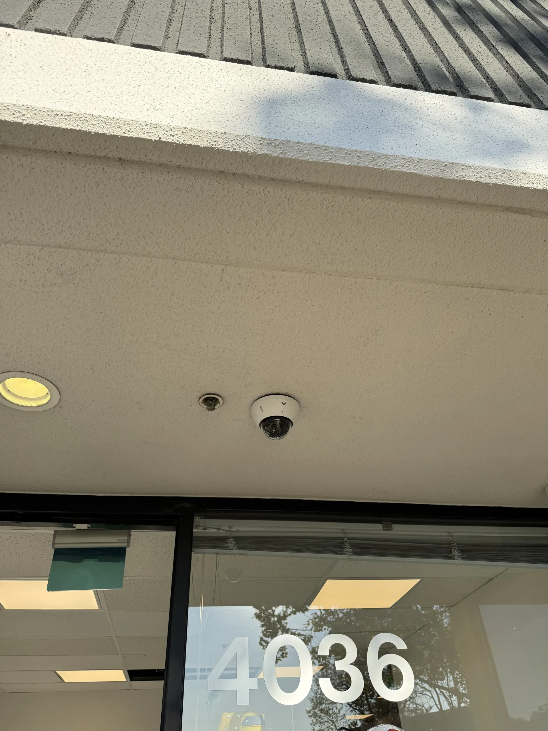 A security dome camera and a small circular sensor mounted on a white exterior ceiling above a glass store entrance.