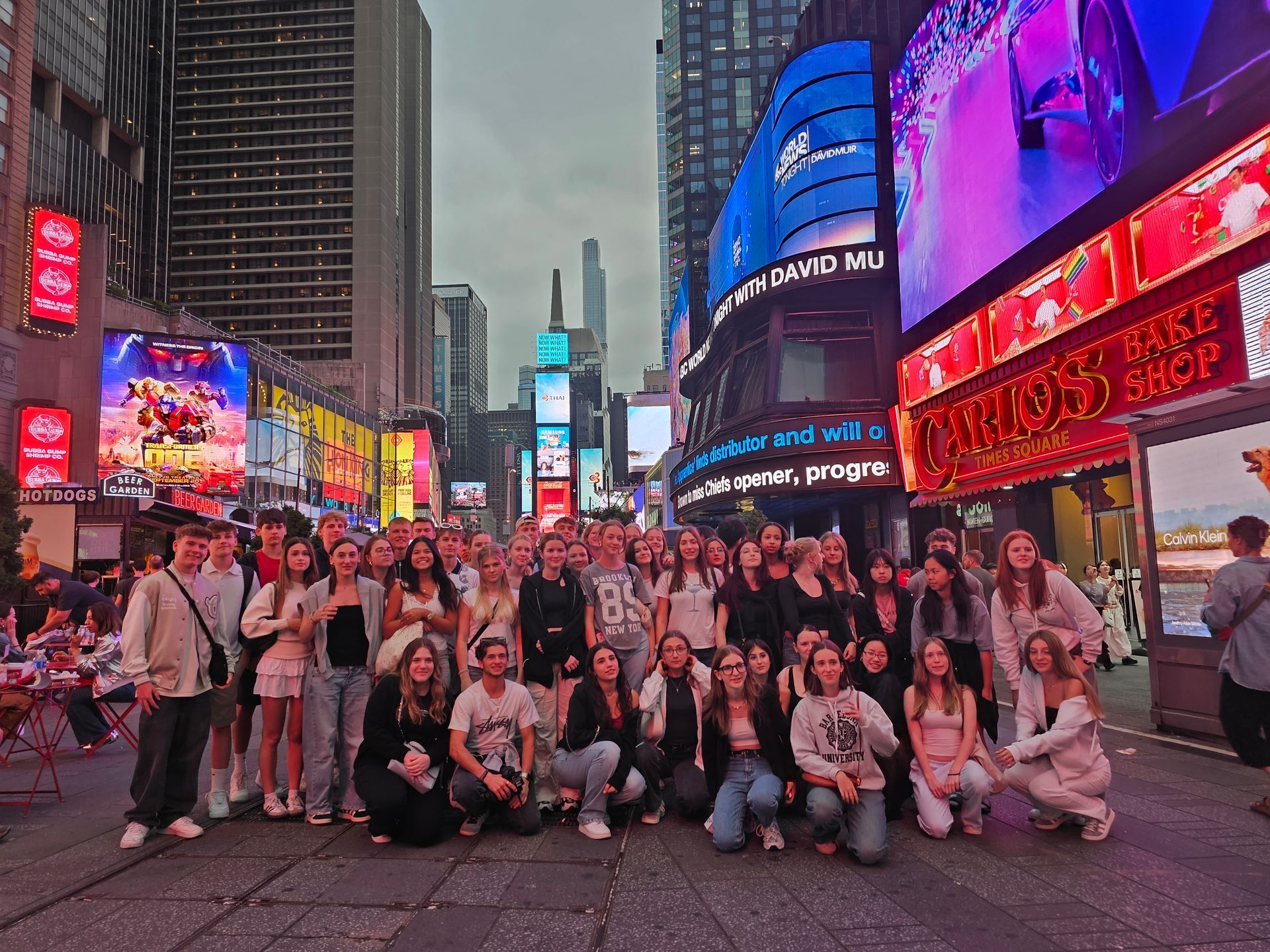Gruppe af mennesker poserer på Times Square, NYC, omgivet af farverige billboards og bygninger i skumringen.