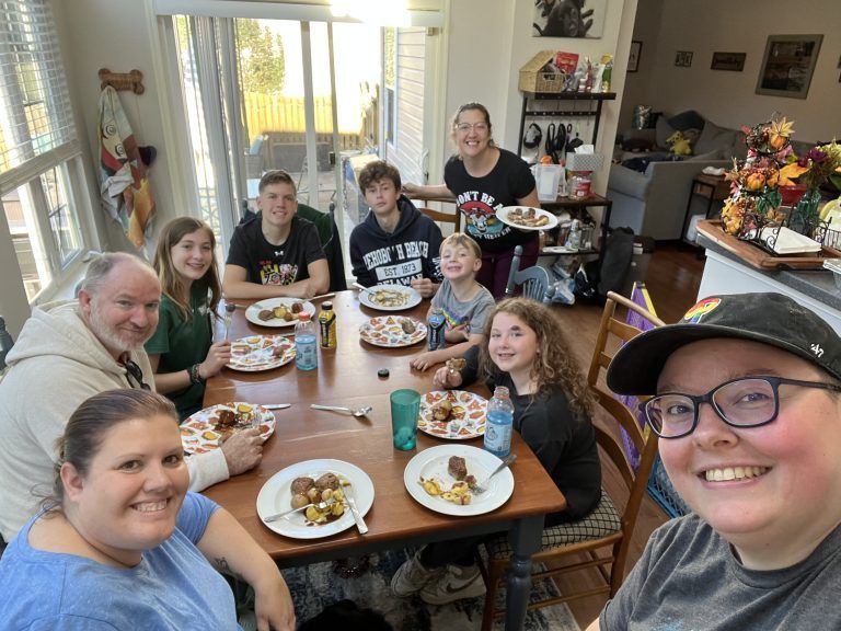 Familie samlet omkring et bord spiser måltid; kvinde tager selfie, sollys fra vinduer.