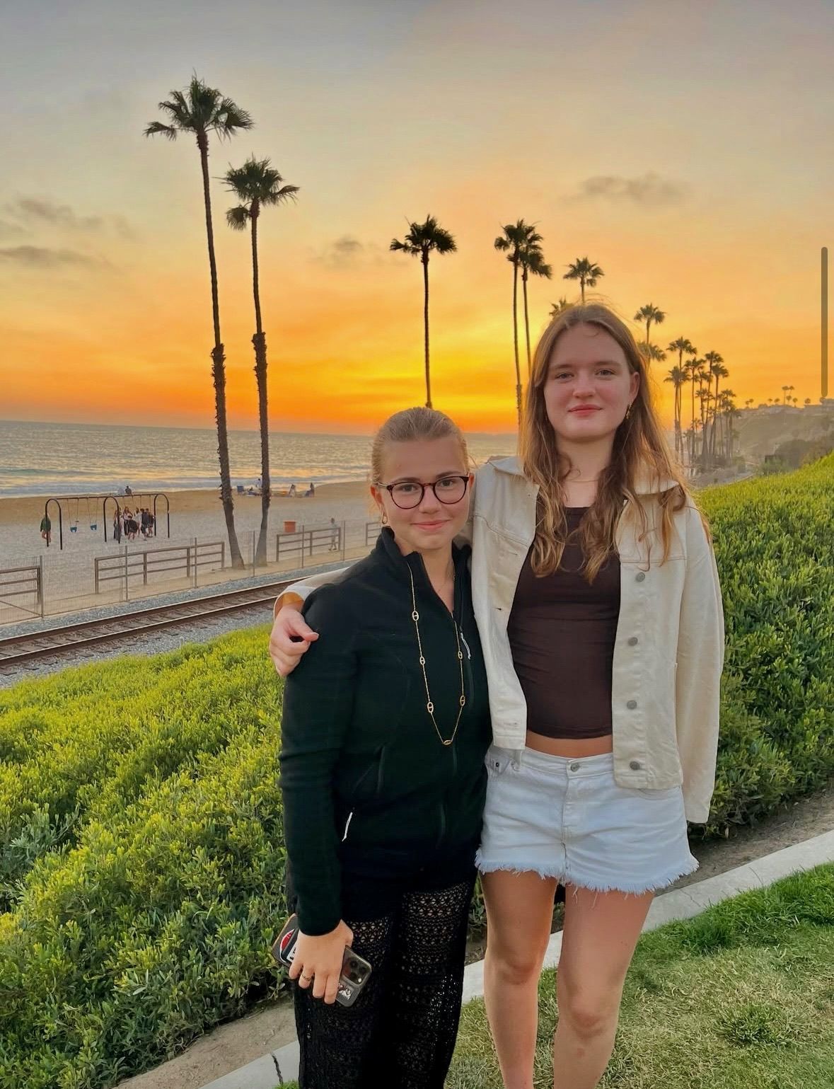 To personer med arme om hinanden poserer nær en strand ved solnedgang. Palmer i baggrunden.