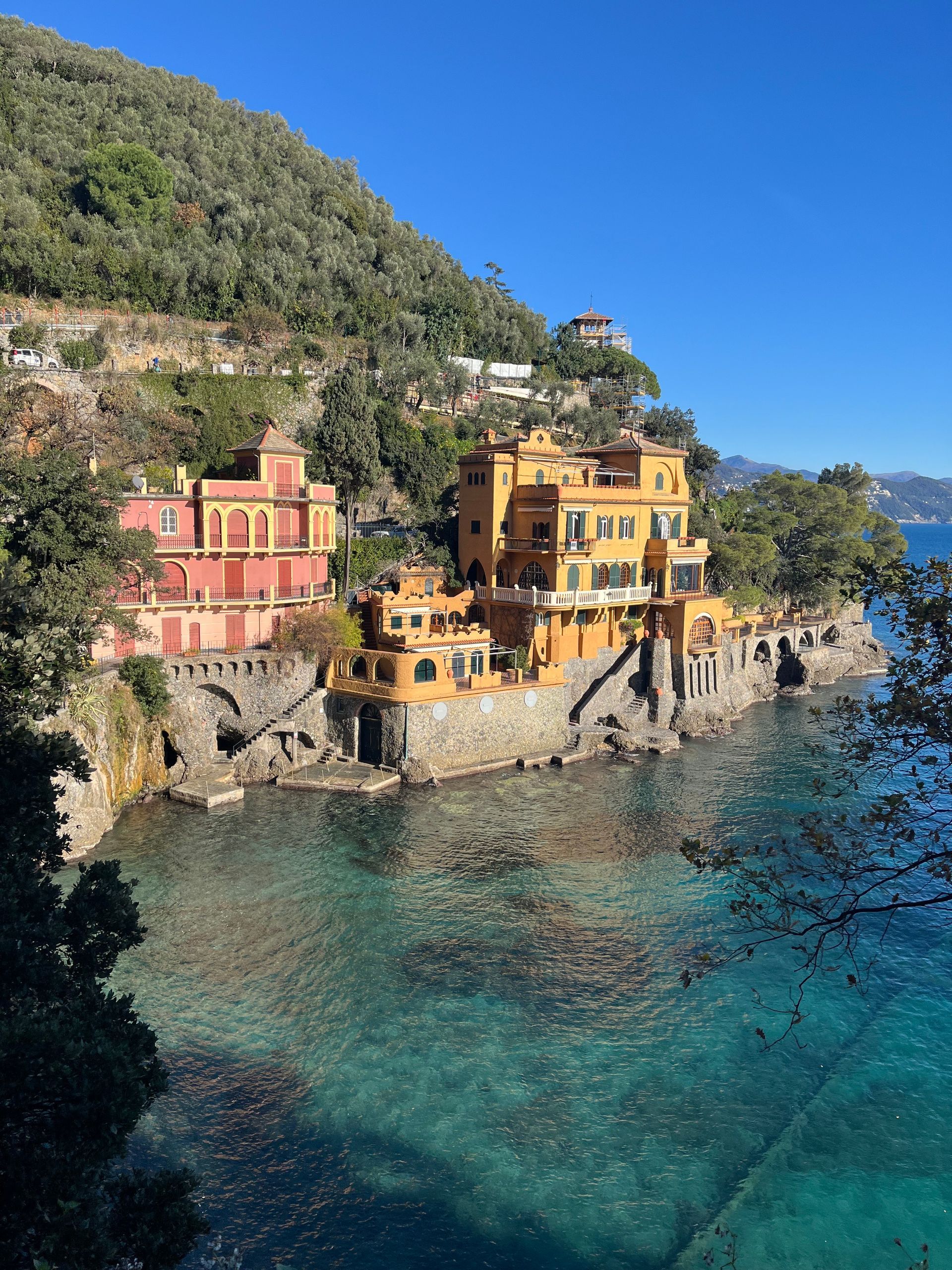 Gule og lyserøde bygninger på en klippeside over turkisblåt vand, med en frodig bjergskråning i baggrunden.