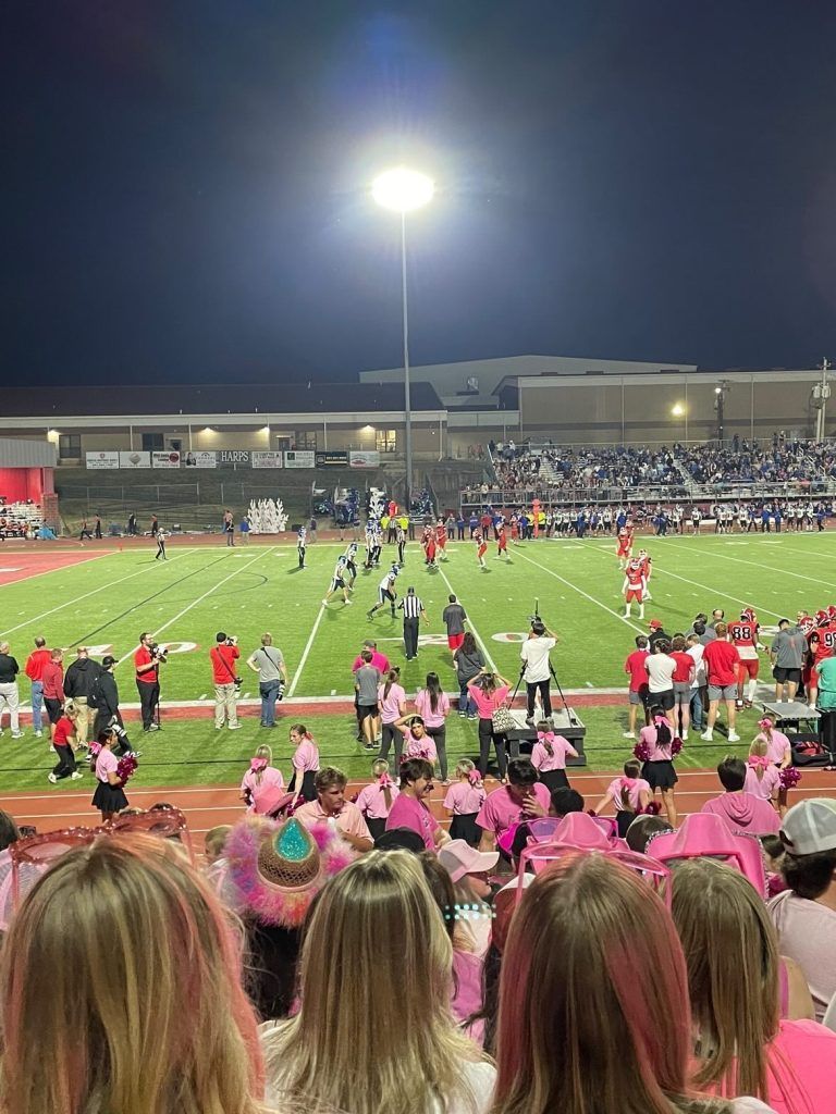 Fodboldkamp om natten under stadionlys; bane, spillere, cheerleaders i lyserødt, publikum på tribunerne.
